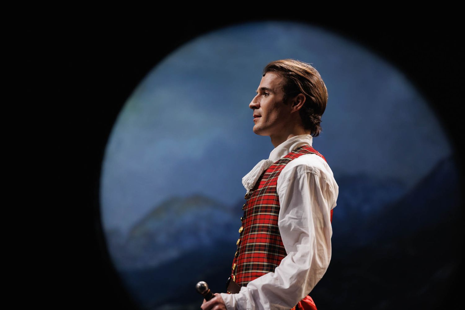 A person in a red plaid vest and white shirt performs on stage with dramatic blue lighting in the background.