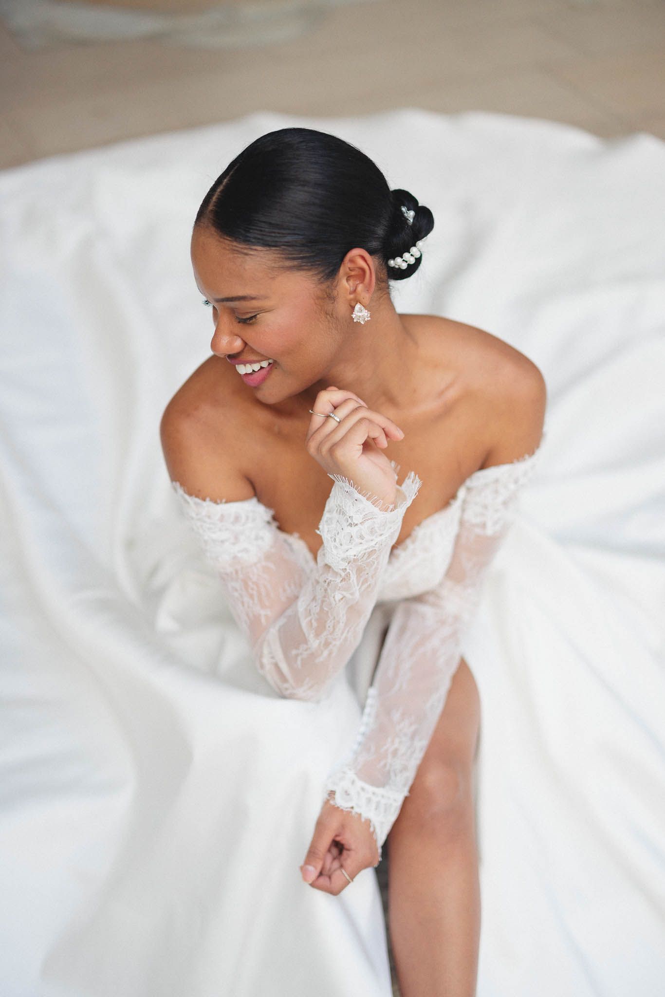 A bride in a white lace wedding dress laughs joyfully while sitting on white bedding.