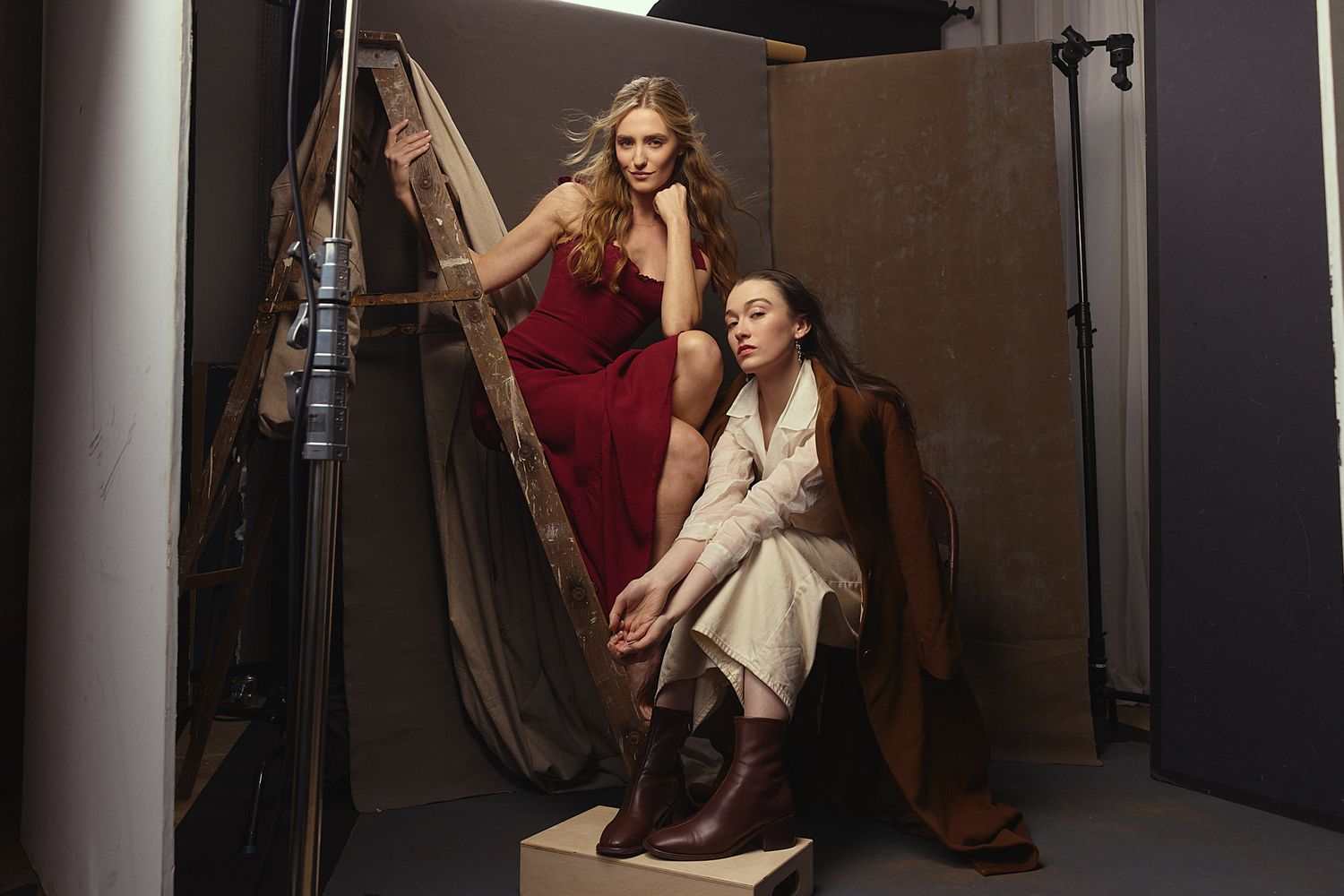 Two models pose in elegant dresses during a fashion photoshoot with studio lighting and brown backdrop.