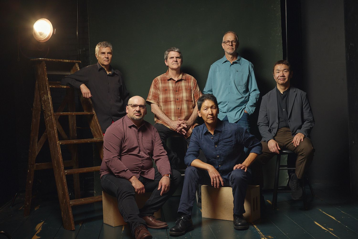 Six men pose for a professional group portrait in a dark studio setting with a wooden ladder prop.