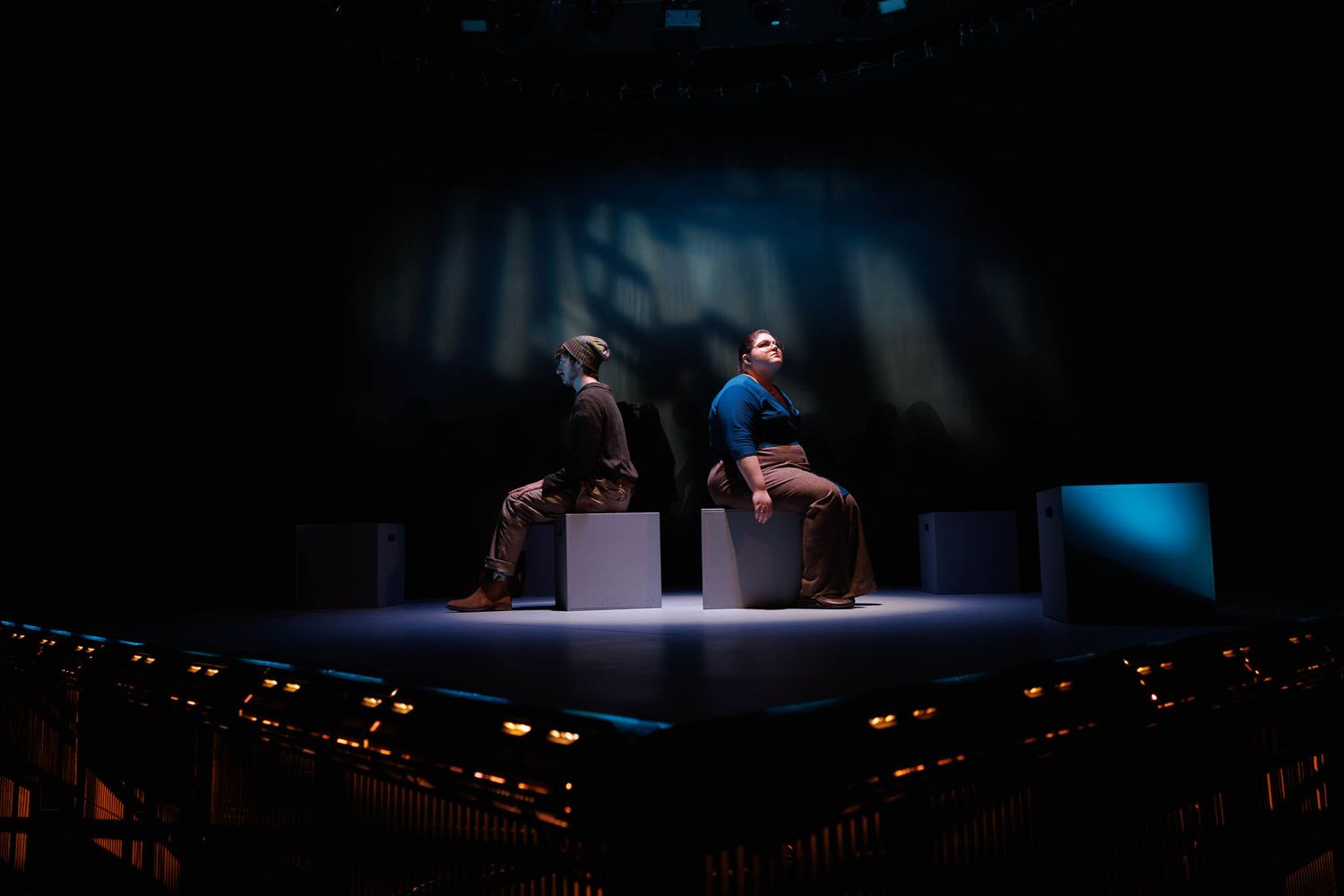 Two performers seated on illuminated blocks on a darkened theater stage.