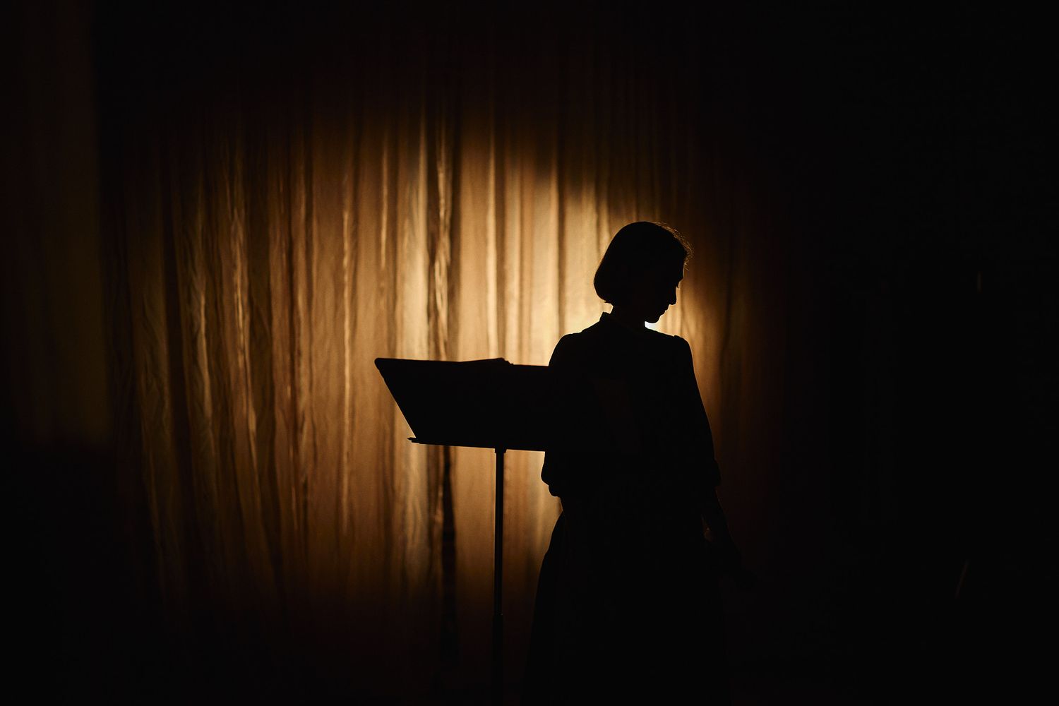 Silhouette of a person reading from an open book or folder, backlit by warm golden curtain light.