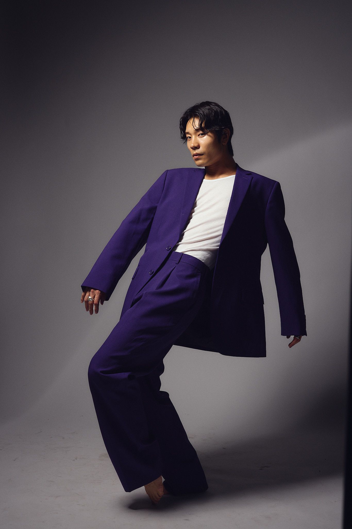 Man in oversized purple suit and white shirt poses dynamically on tiptoe against gray studio background.
