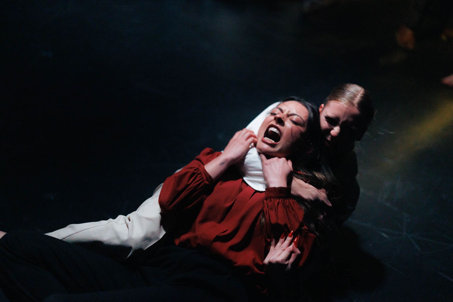 A performer in a red costume passionately acts out an emotional scene on a dark stage during a theatrical performance.