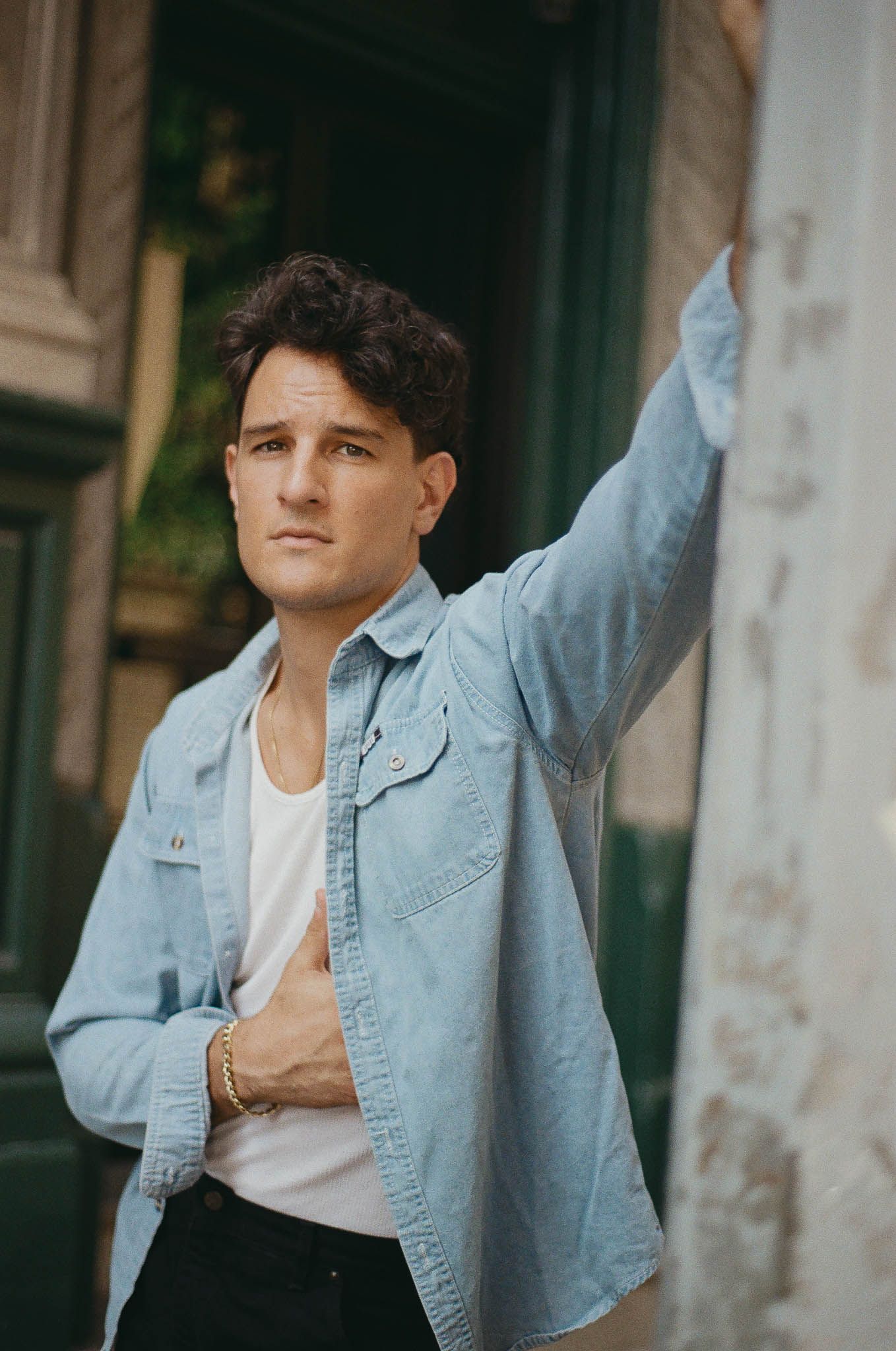 A person in a light denim jacket and white t-shirt poses casually against a wall in an outdoor setting.