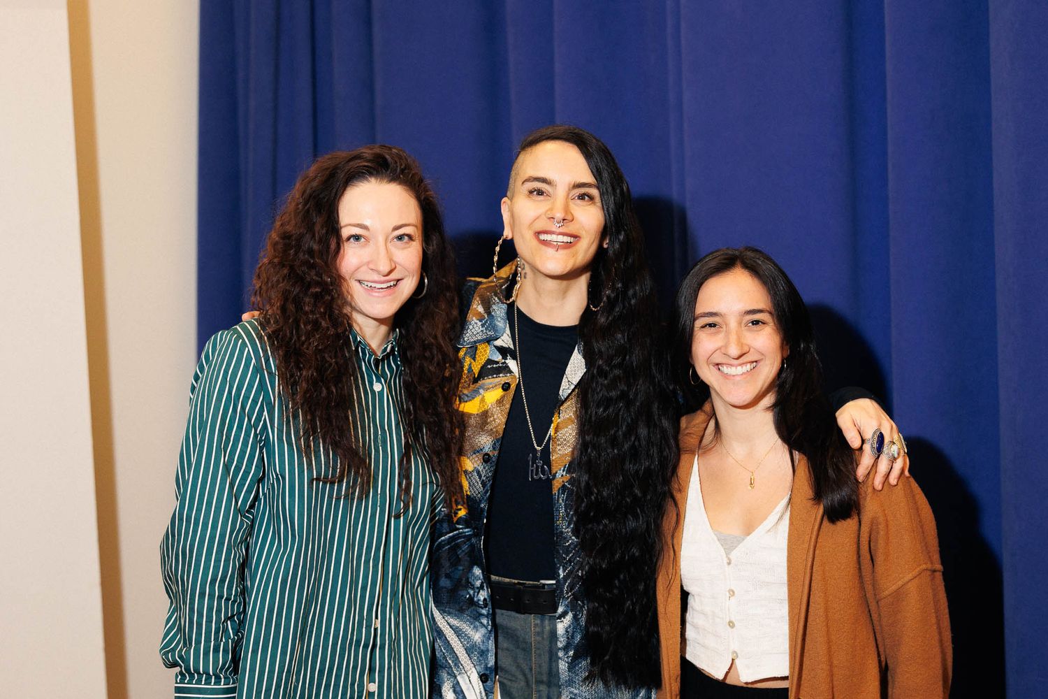 Three women pose together against a blue backdrop wearing casual clothing.