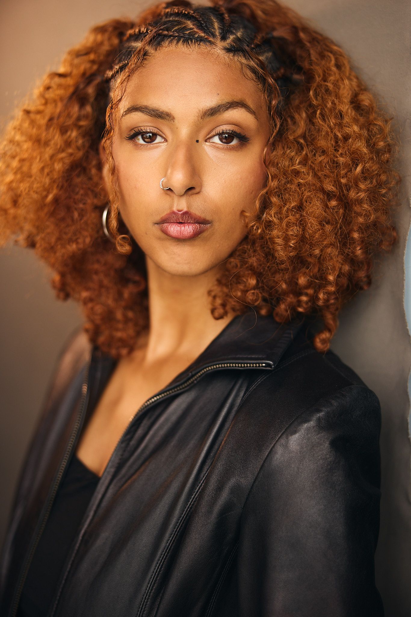 Young woman with curly auburn hair, nose ring, and hoop earrings wearing a black leather jacket.
