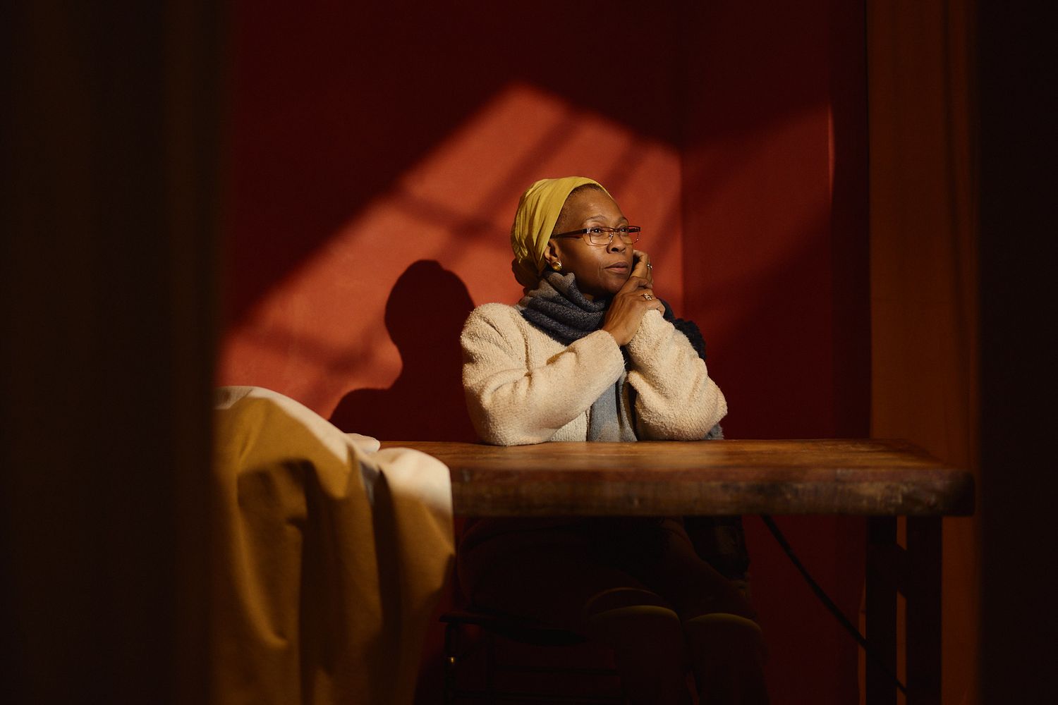 A woman in a yellow headscarf sits thoughtfully at a wooden table, bathed in warm red light and shadows.