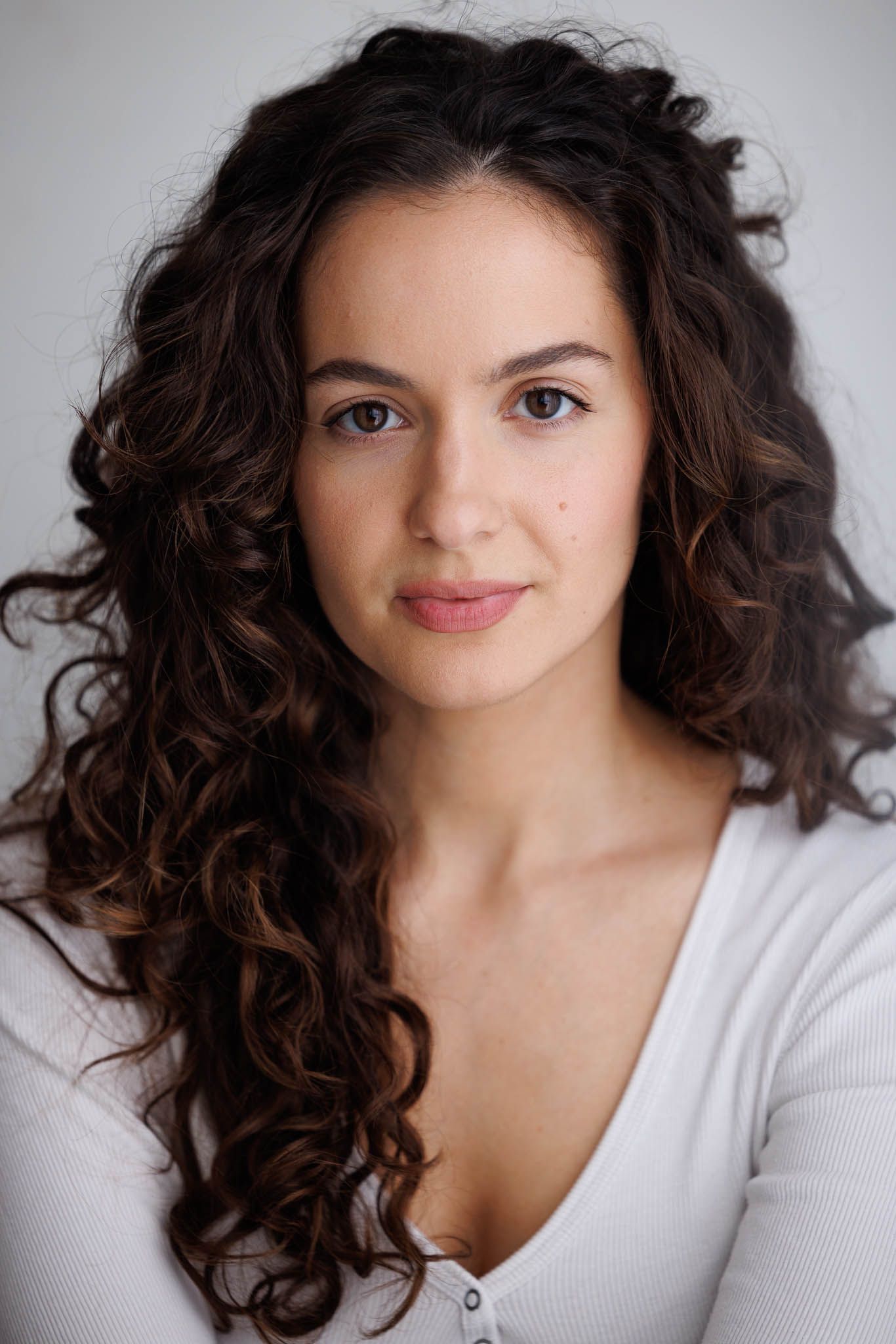 Professional headshot of a young woman with long curly dark hair wearing a white top against a light background.