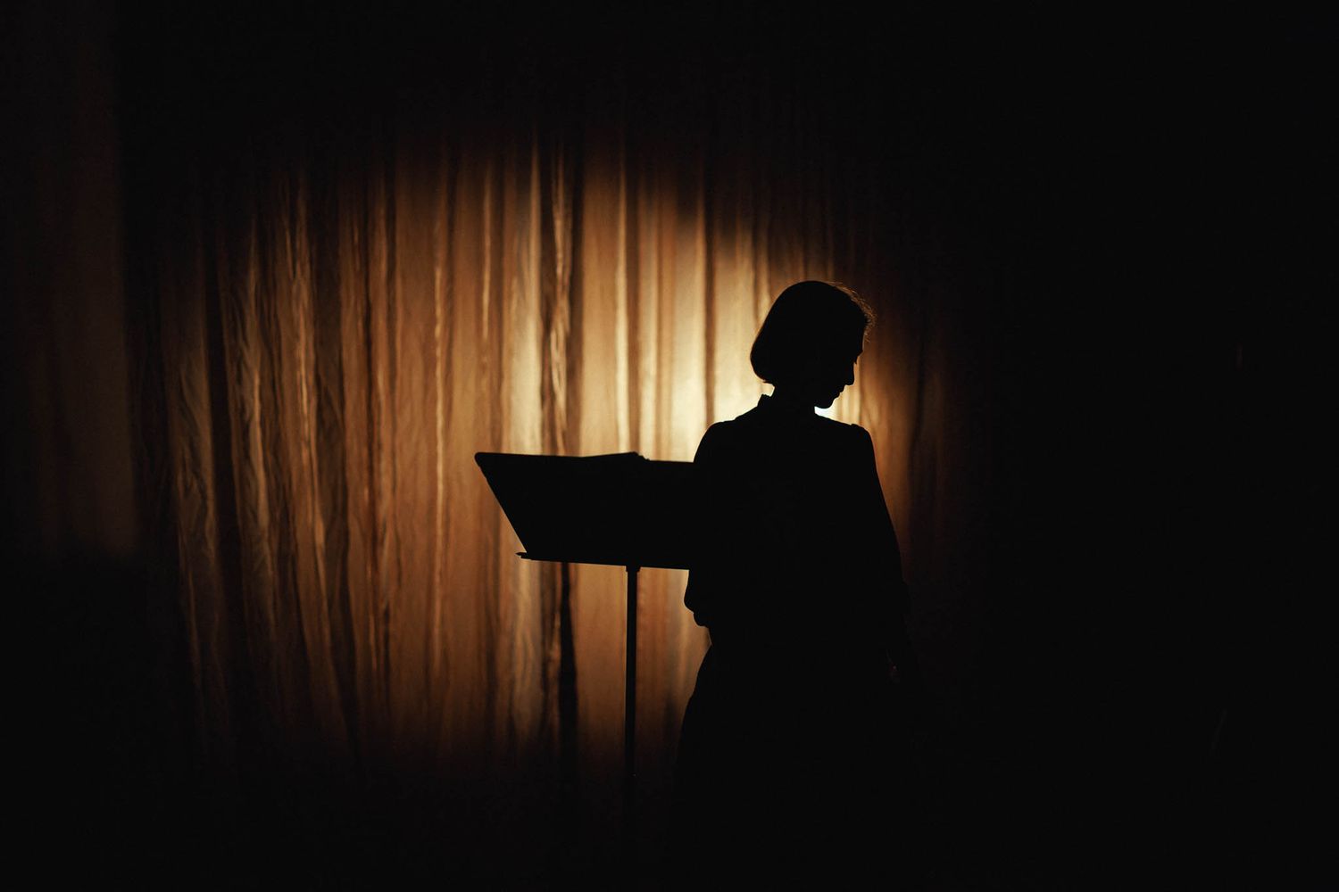 A series of silhouetted figures against a warm backlit wooden wall creates a dramatic theatrical scene.