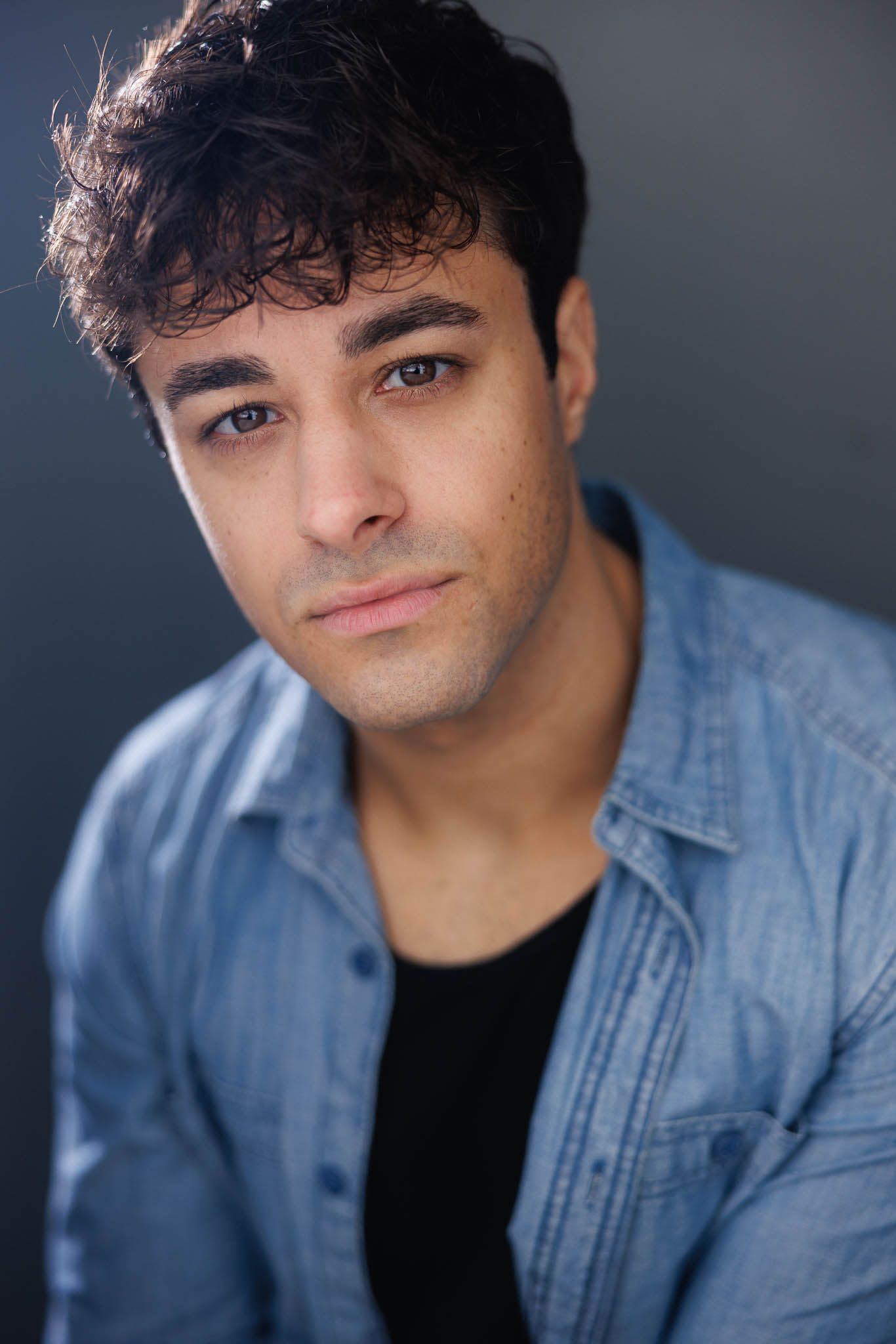 Professional headshot portrait against dark background wearing denim shirt and black t-shirt.