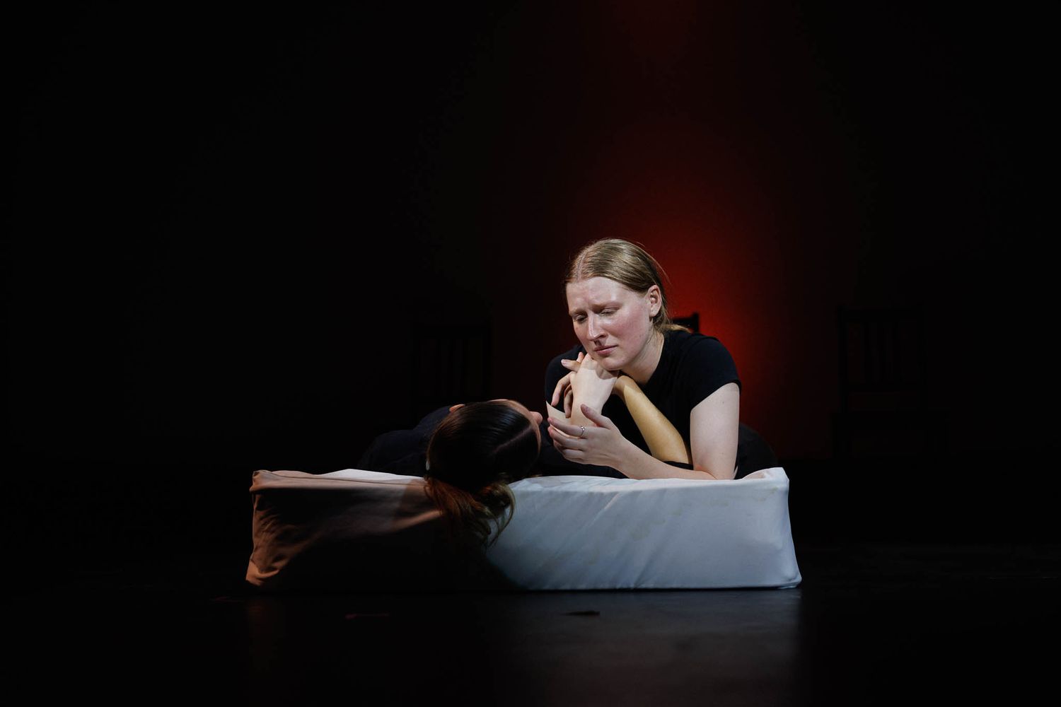 A dramatic theater scene with performer resting on white cushions under moody lighting.