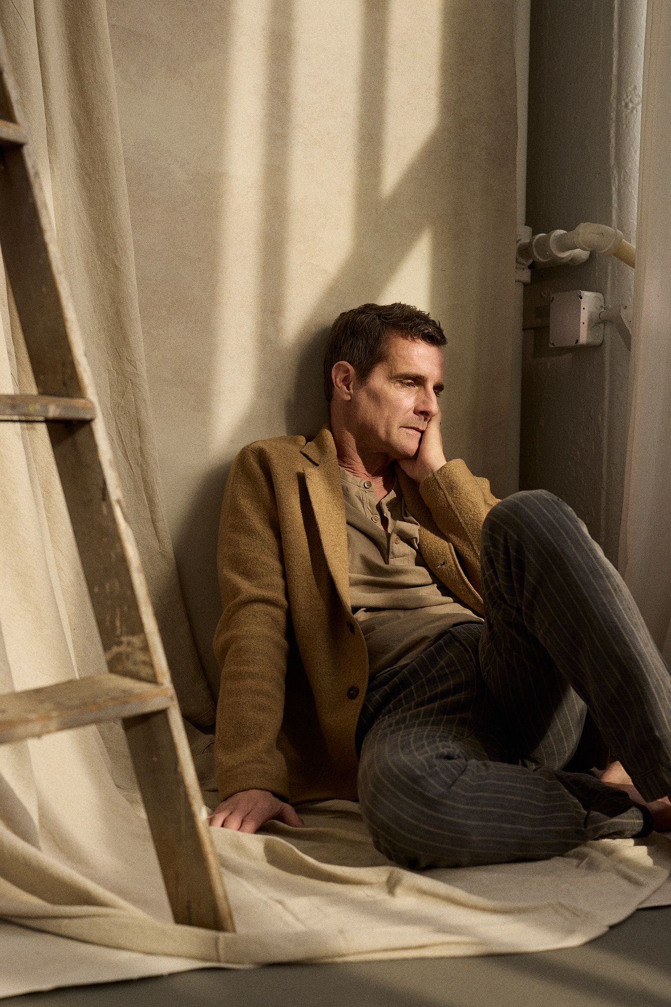 A contemplative figure sits on the floor against a wall in warm lighting wearing a brown jacket.