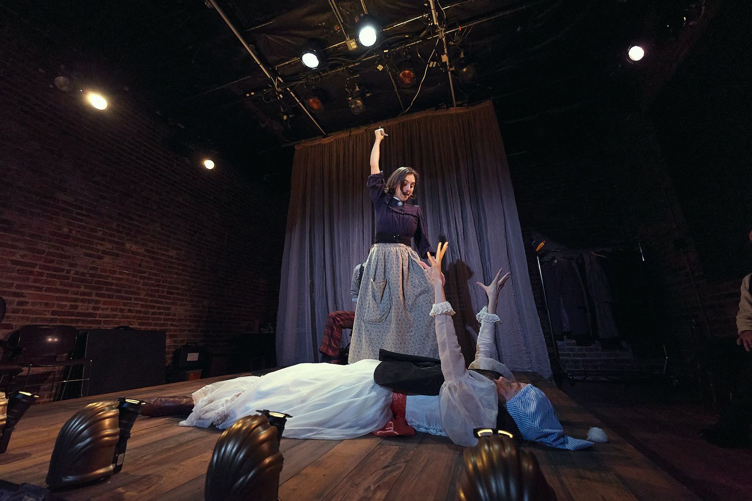 Theatrical performance with actress standing triumphantly over fallen actor on stage with brick walls and dramatic lighting.