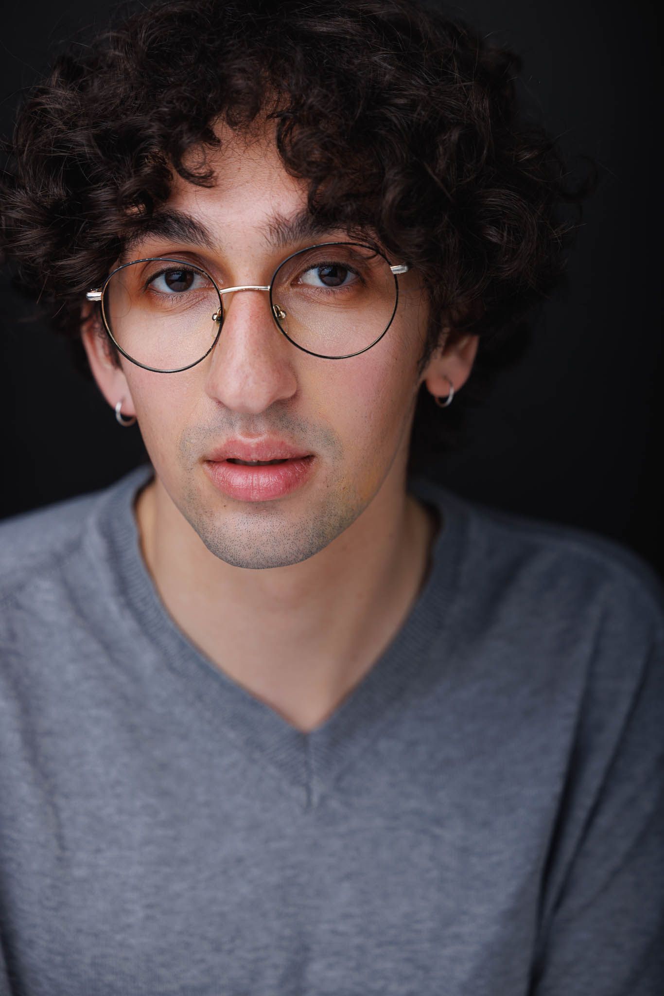 Portrait of person wearing round glasses and gray v-neck shirt against dark background.