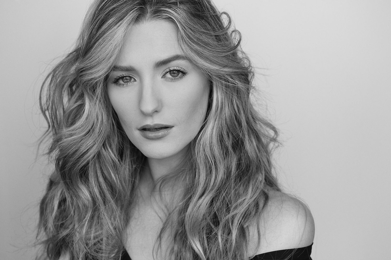 Black and white portrait of a woman with wavy blonde hair and dark lipstick wearing an off-shoulder top.