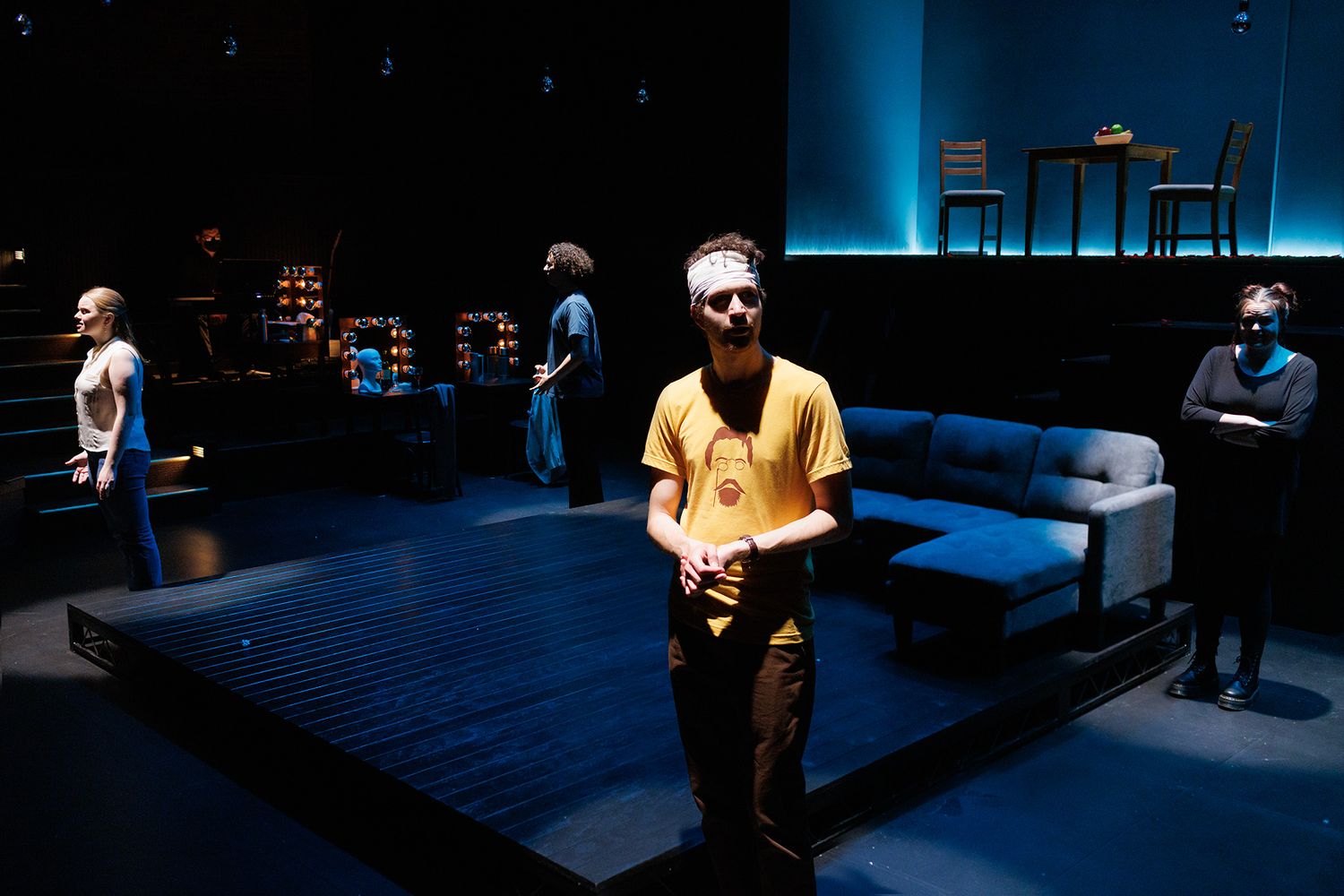 Stage performance featuring actors in a minimalist blue-lit set with furniture.