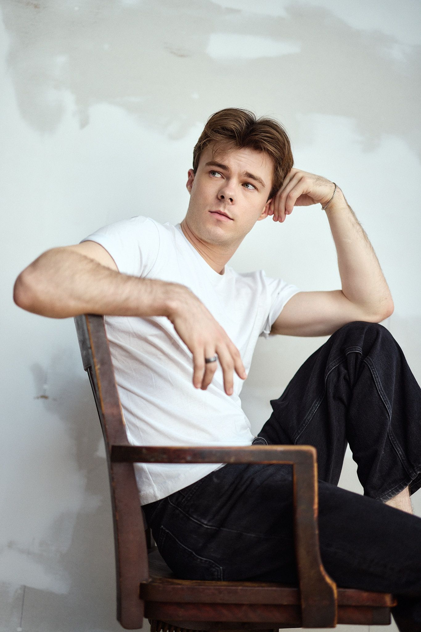 Young man in white t-shirt and black jeans sits relaxed on wooden chair against white wall.