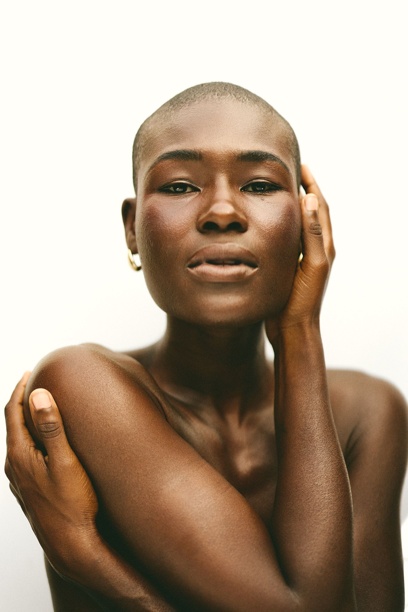 Bald Black woman with glowing skin poses gracefully, hand touching face, wearing gold earring on white background.
