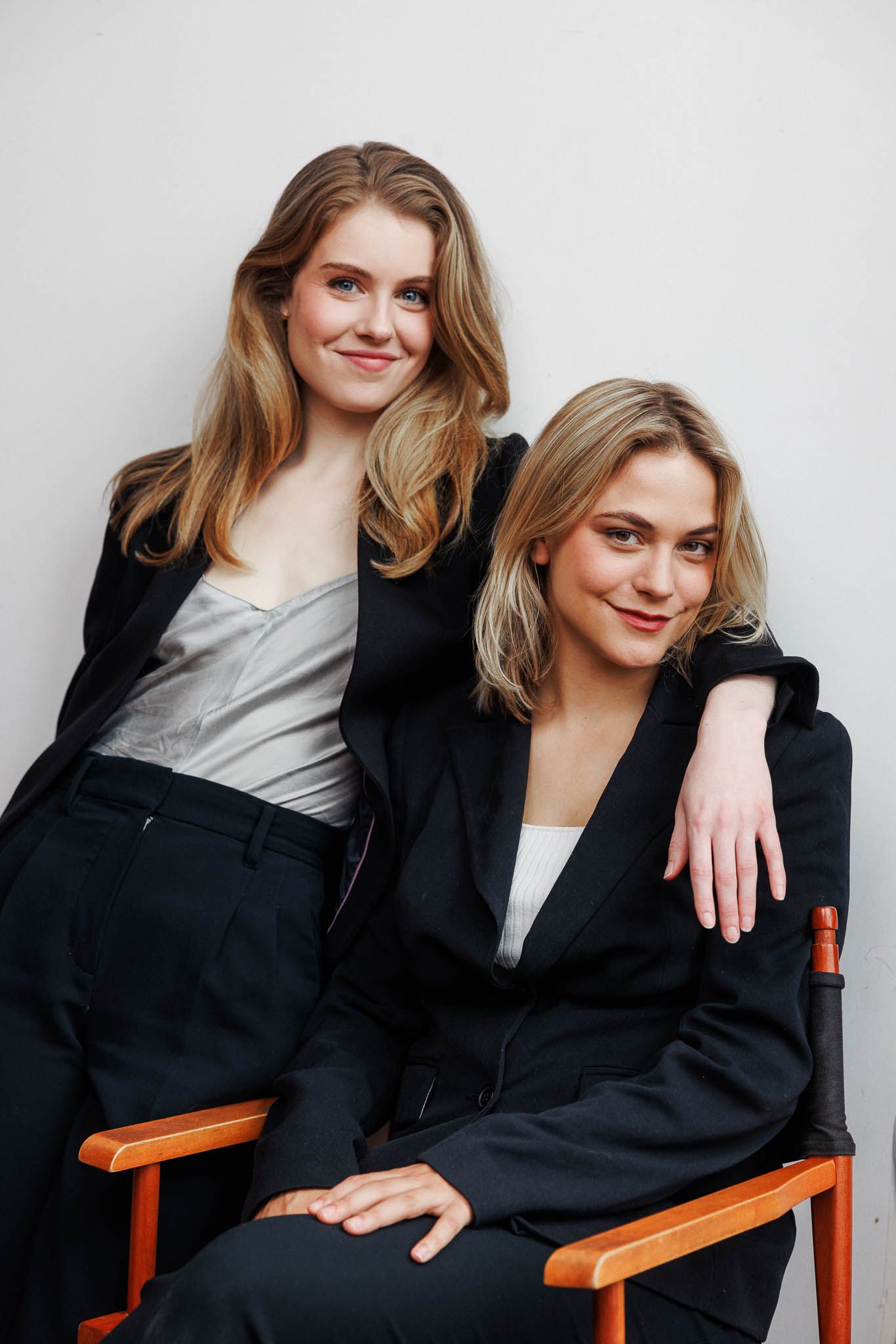 Professional studio portrait of two people in black blazers sitting on wooden chair.