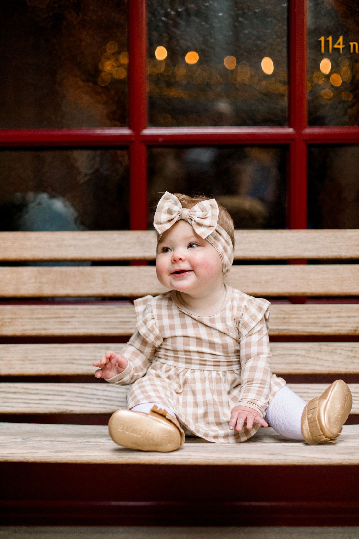 Smiling baby girl in plaid dress and bow headband sitting on wooden bench near red-framed window.
