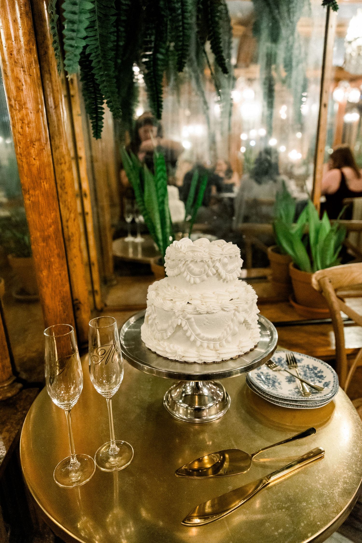 Two-tier white wedding cake on gold stand with champagne flutes and utensils, surrounded by ferns and mirrors.