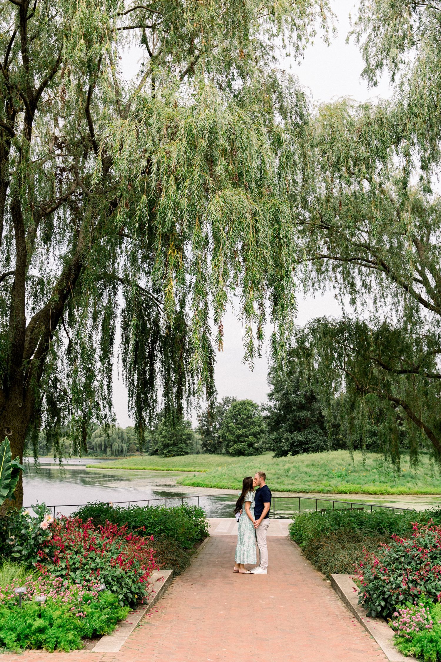 Couple shares a kiss on a brick pathway beneath a weeping willow tree beside a serene garden pond.