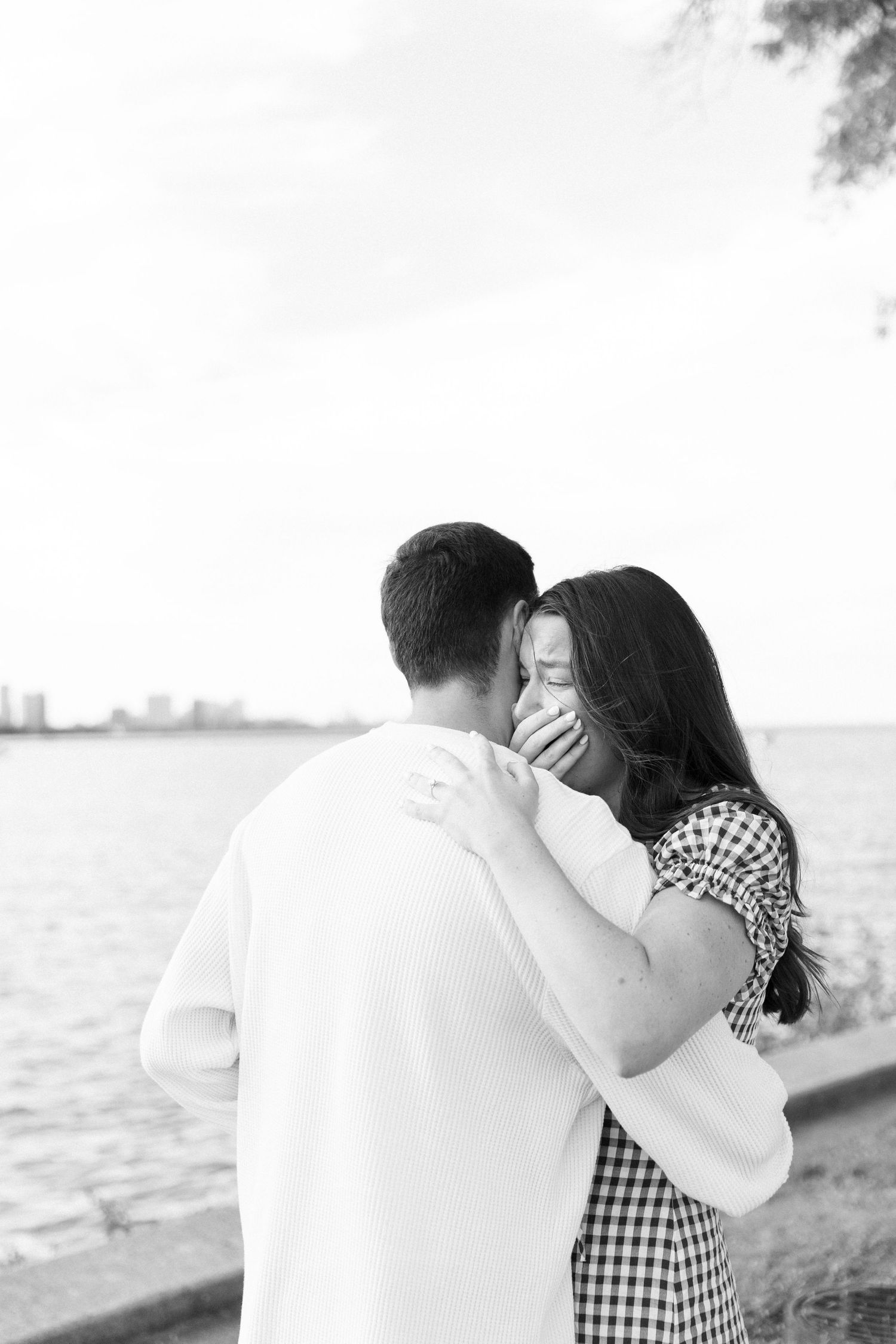 Black and white photo of a couple embracing near a waterfront, woman covering her mouth in surprise.
