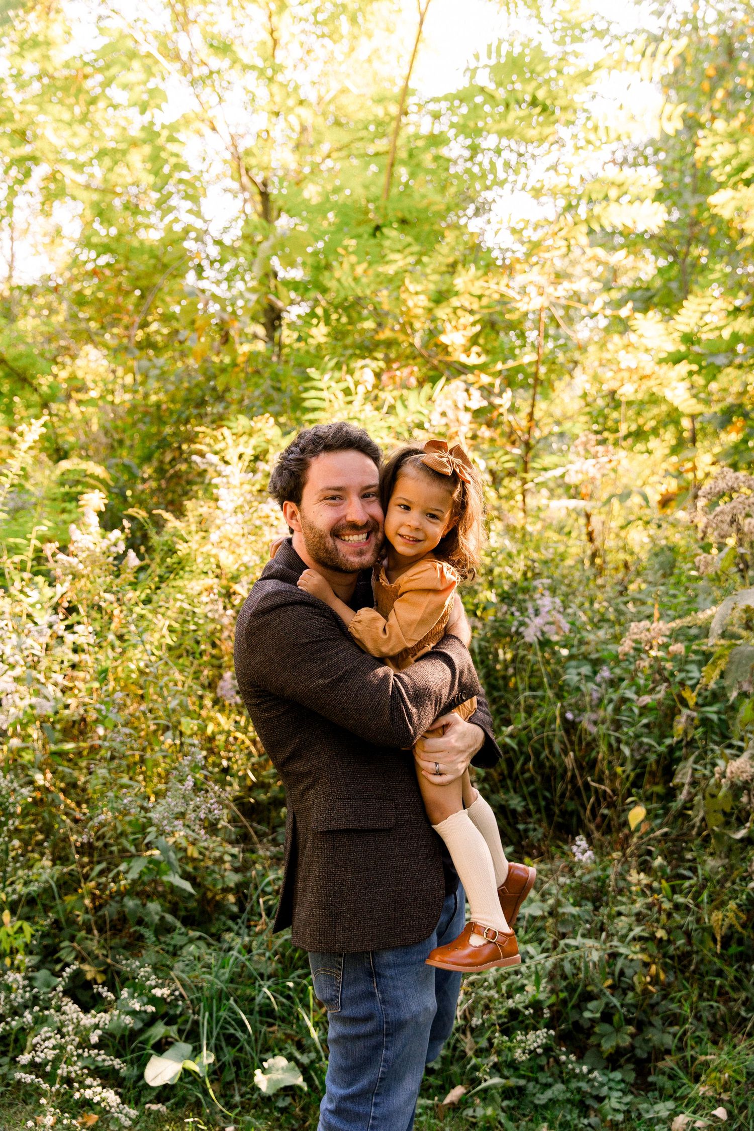A smiling man holds a young child in a sunlit forest setting during autumn.