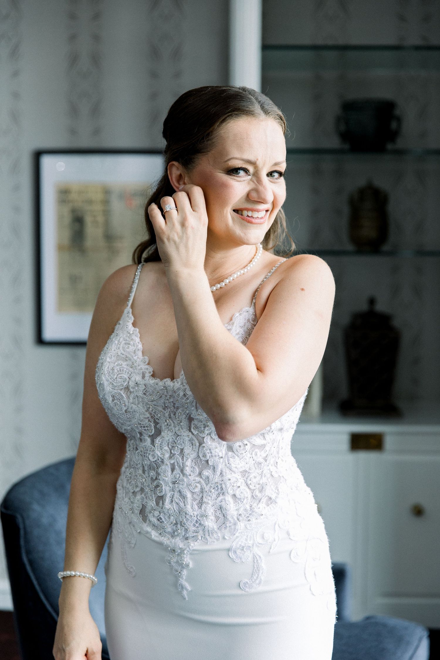 Smiling bride in lace wedding gown adjusting pearl earring, wearing pearl necklace and bracelet indoors.