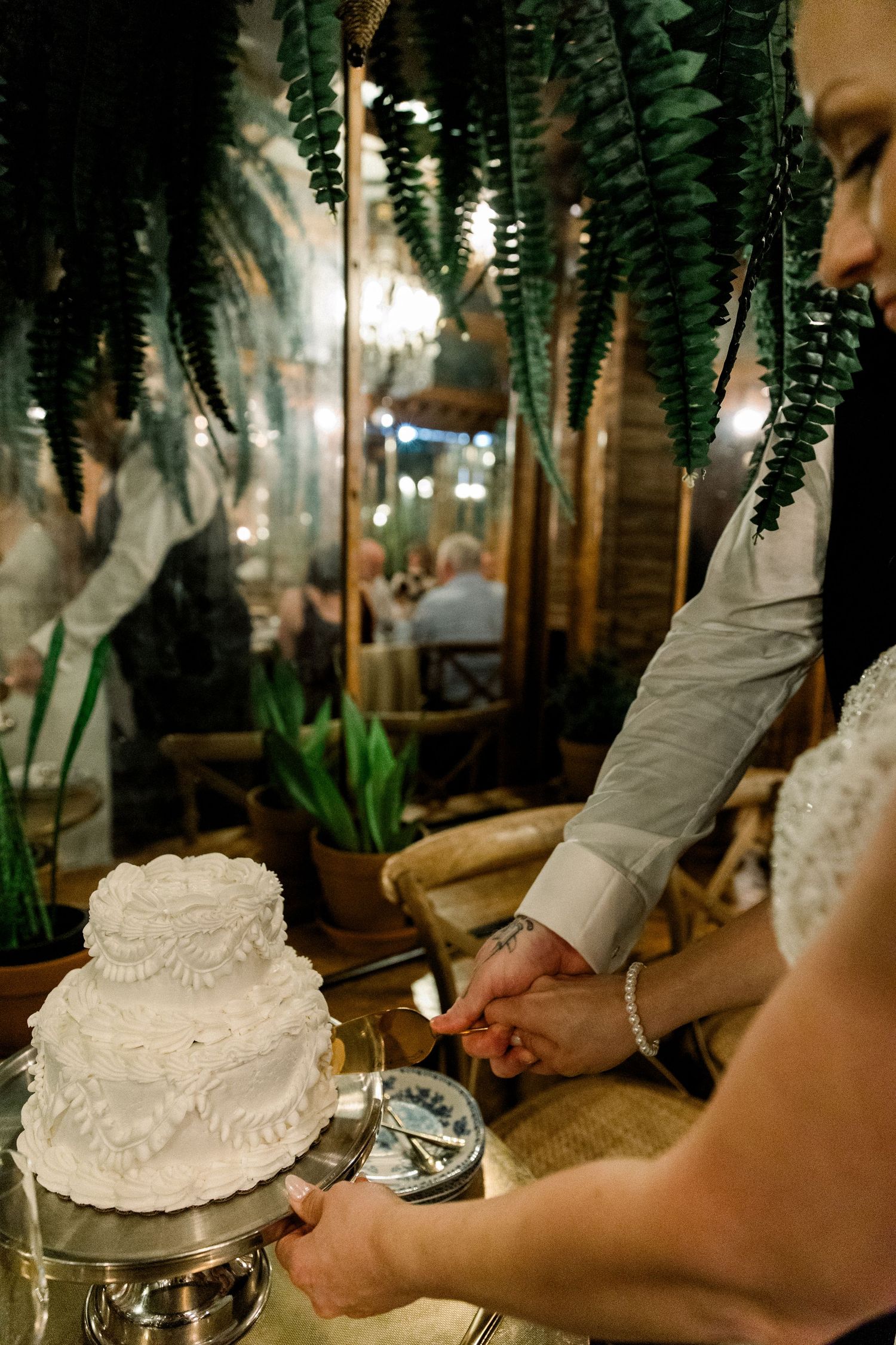 Bride and groom cutting two-tier white wedding cake surrounded by hanging ferns.