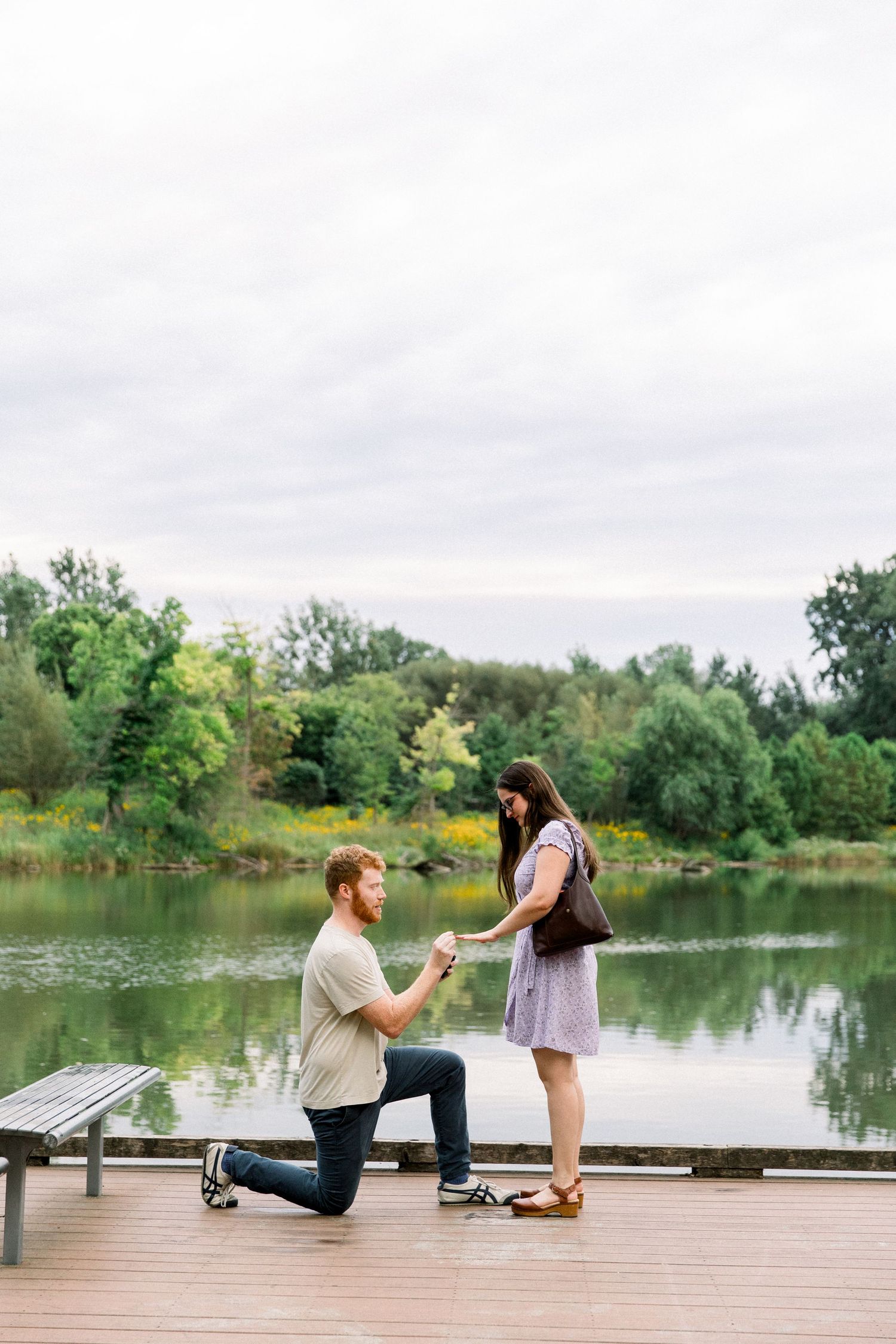 Man kneeling on one knee proposing to woman on wooden dock by serene lake surrounded by lush green trees.