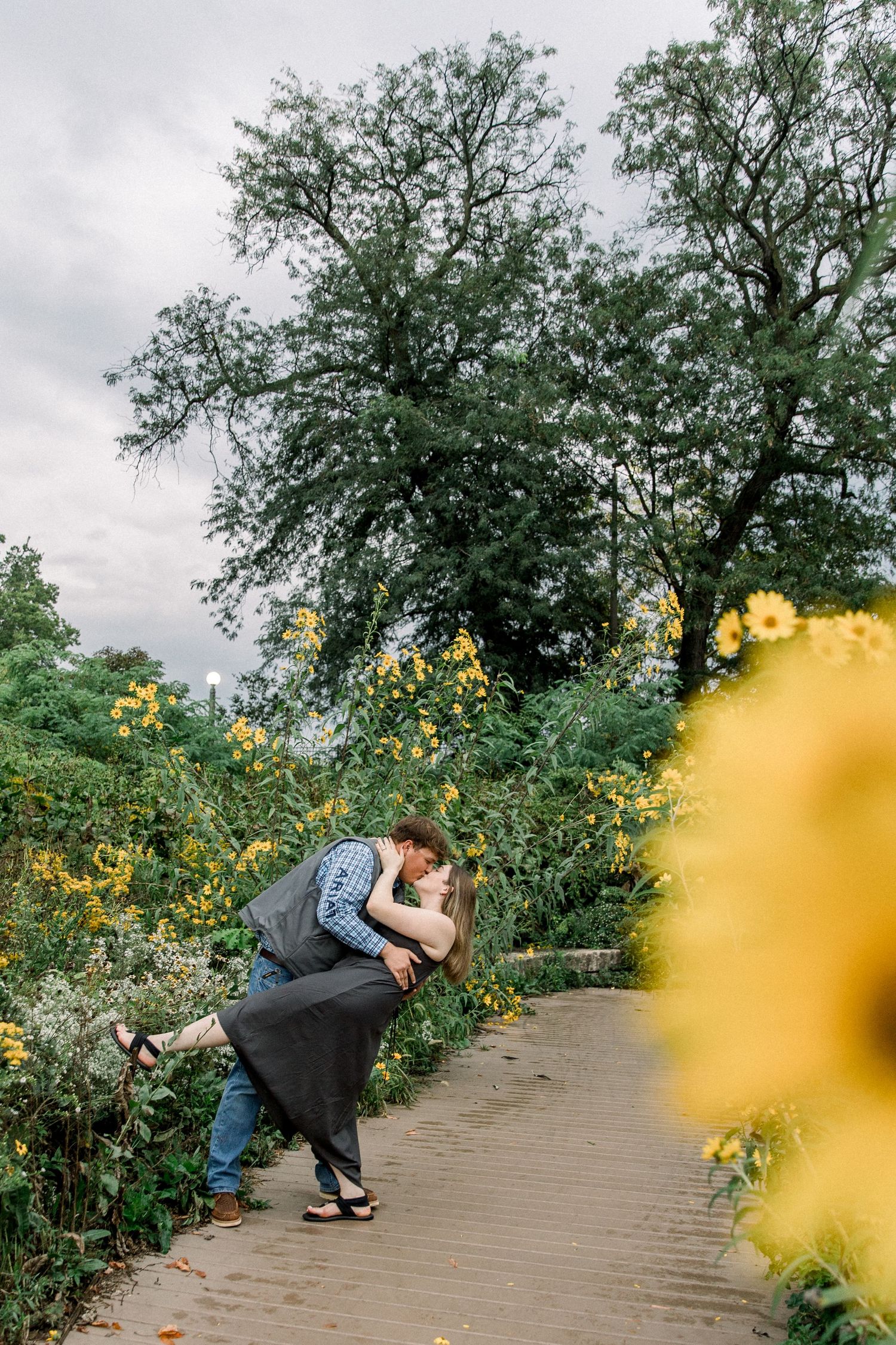 Couple sharing a romantic dip kiss on a garden path surrounded by yellow wildflowers and lush green trees.