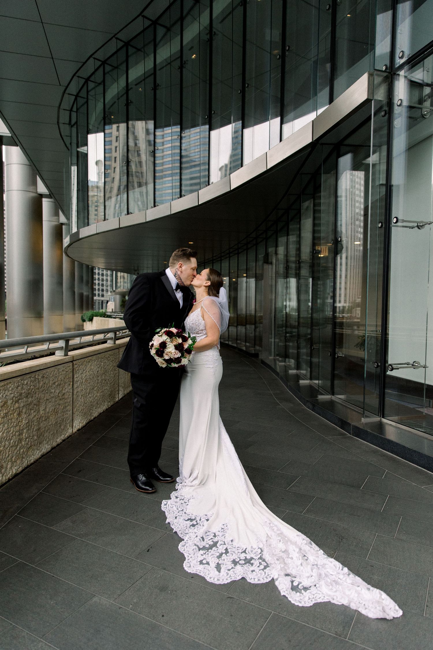 Bride and groom share a kiss outside a modern glass building, her long lace cathedral veil trailing elegantly.