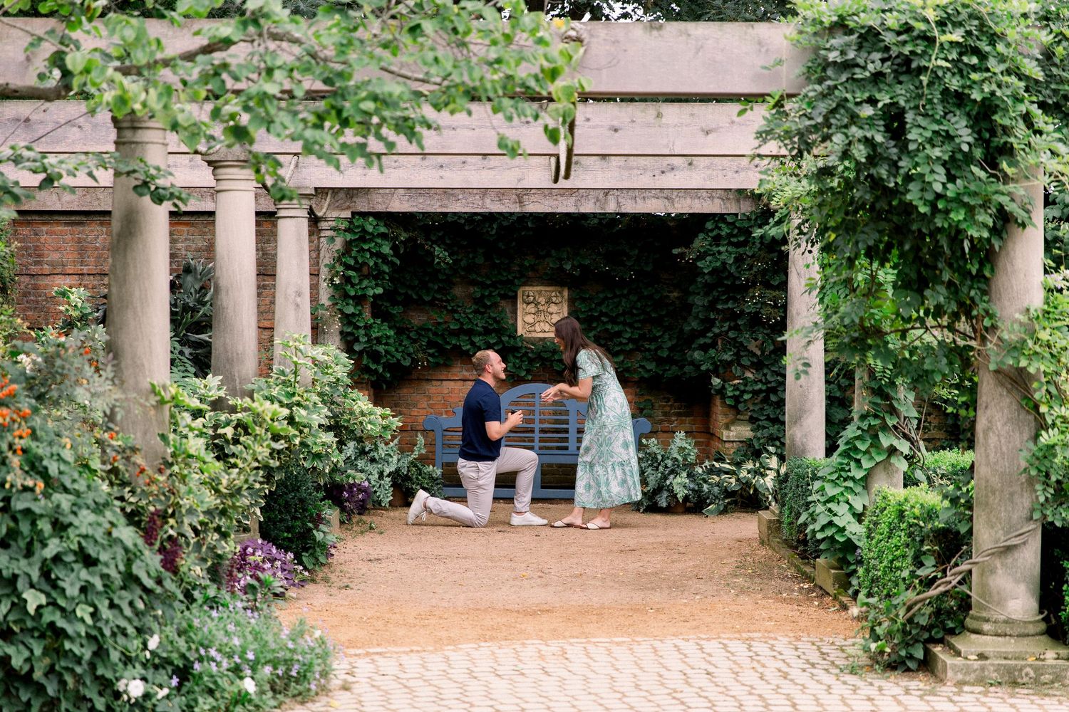 Man proposes to woman on one knee in a lush garden pergola surrounded by greenery and flowers.