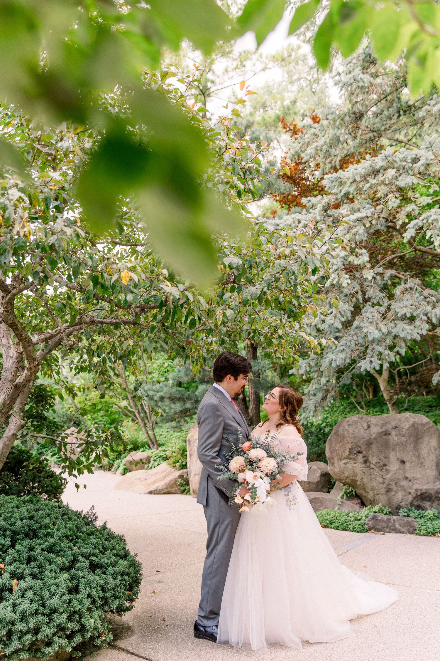 A bride and groom share a romantic moment in a lush garden, surrounded by blooming trees, as she holds a soft floral bouquet.