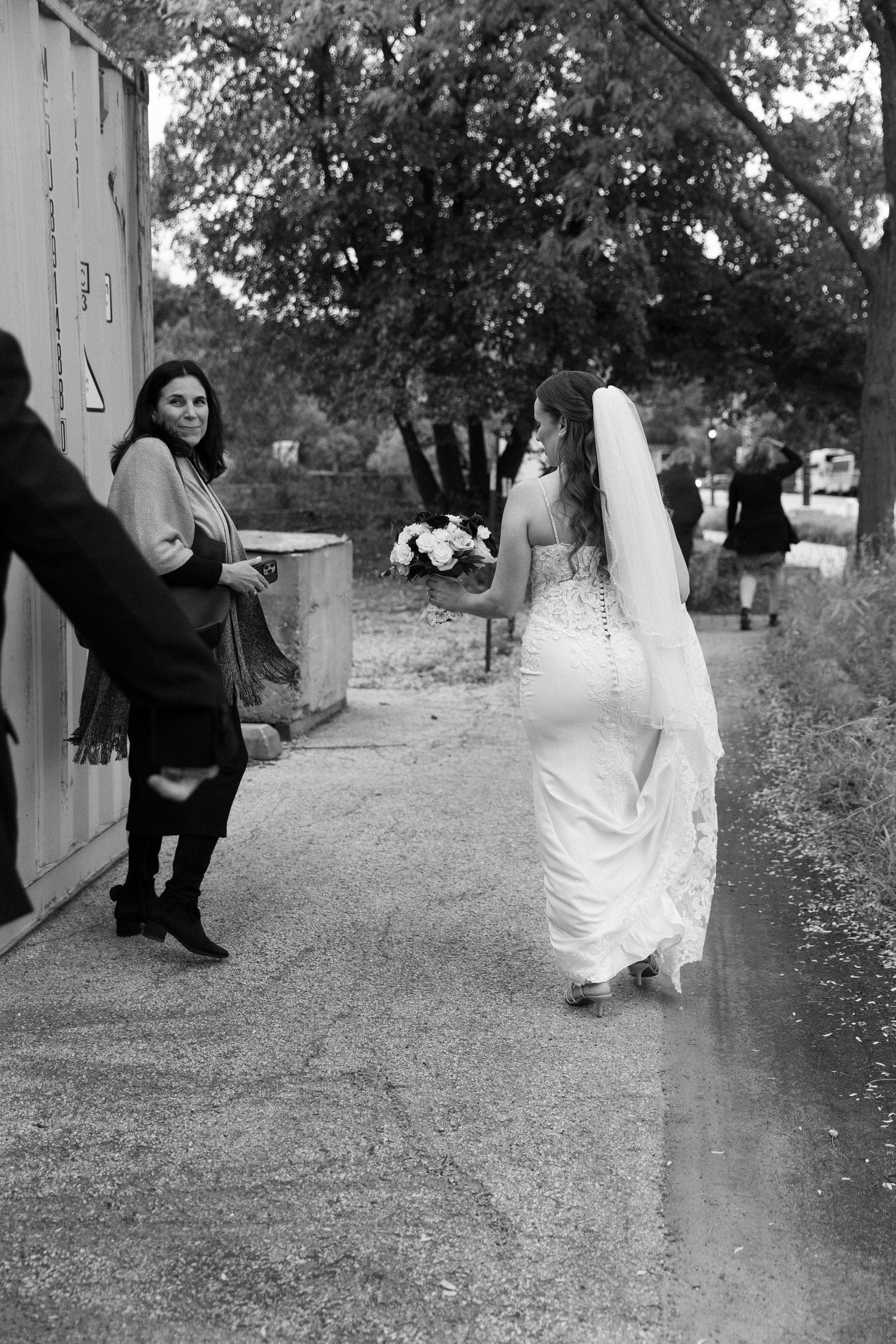 Black and white photo of a bride in a white gown holding a bouquet, walking past a smiling woman on a sidewalk.