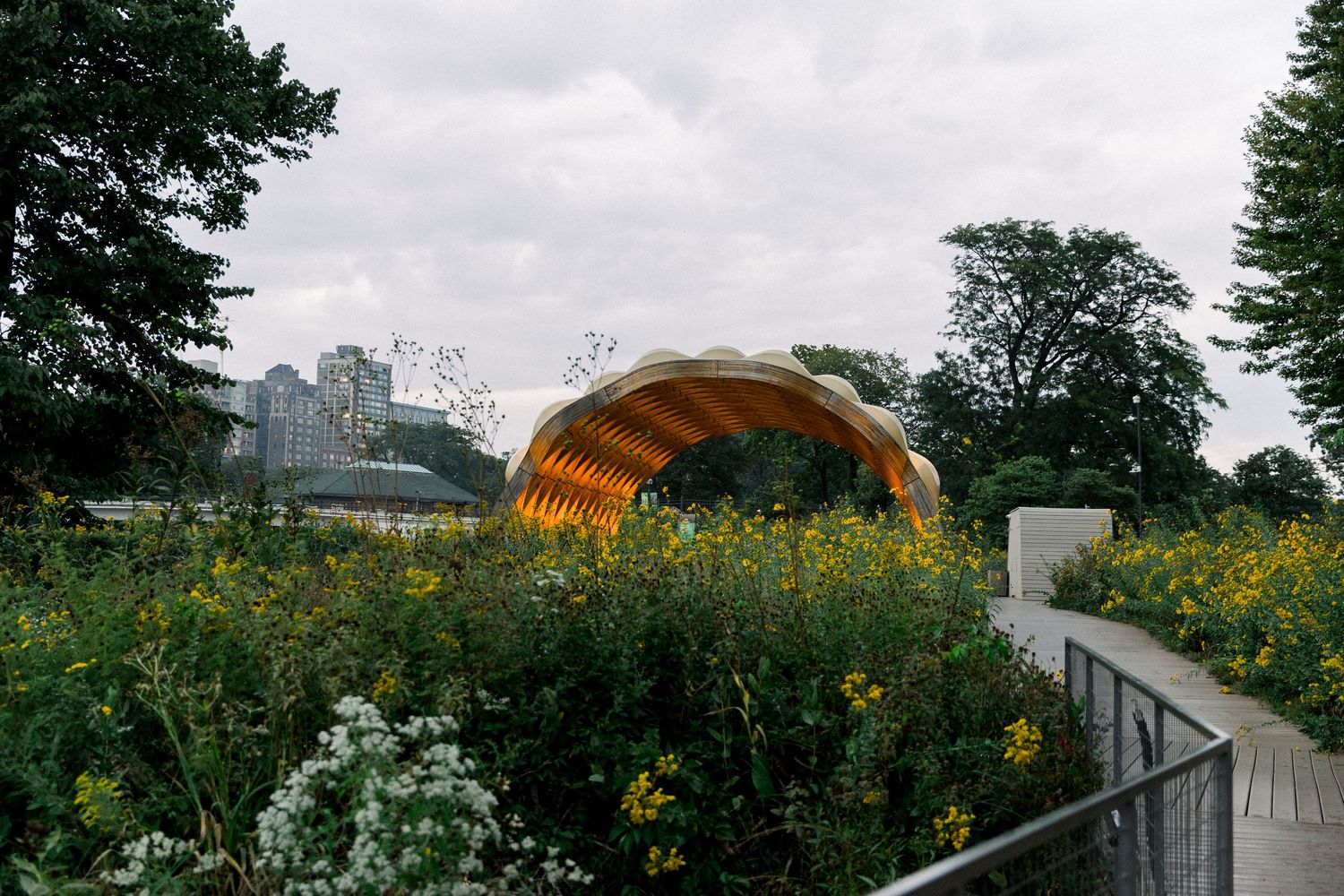 Golden shell-shaped pavilion surrounded by wildflowers and lush greenery in a scenic park under overcast skies.