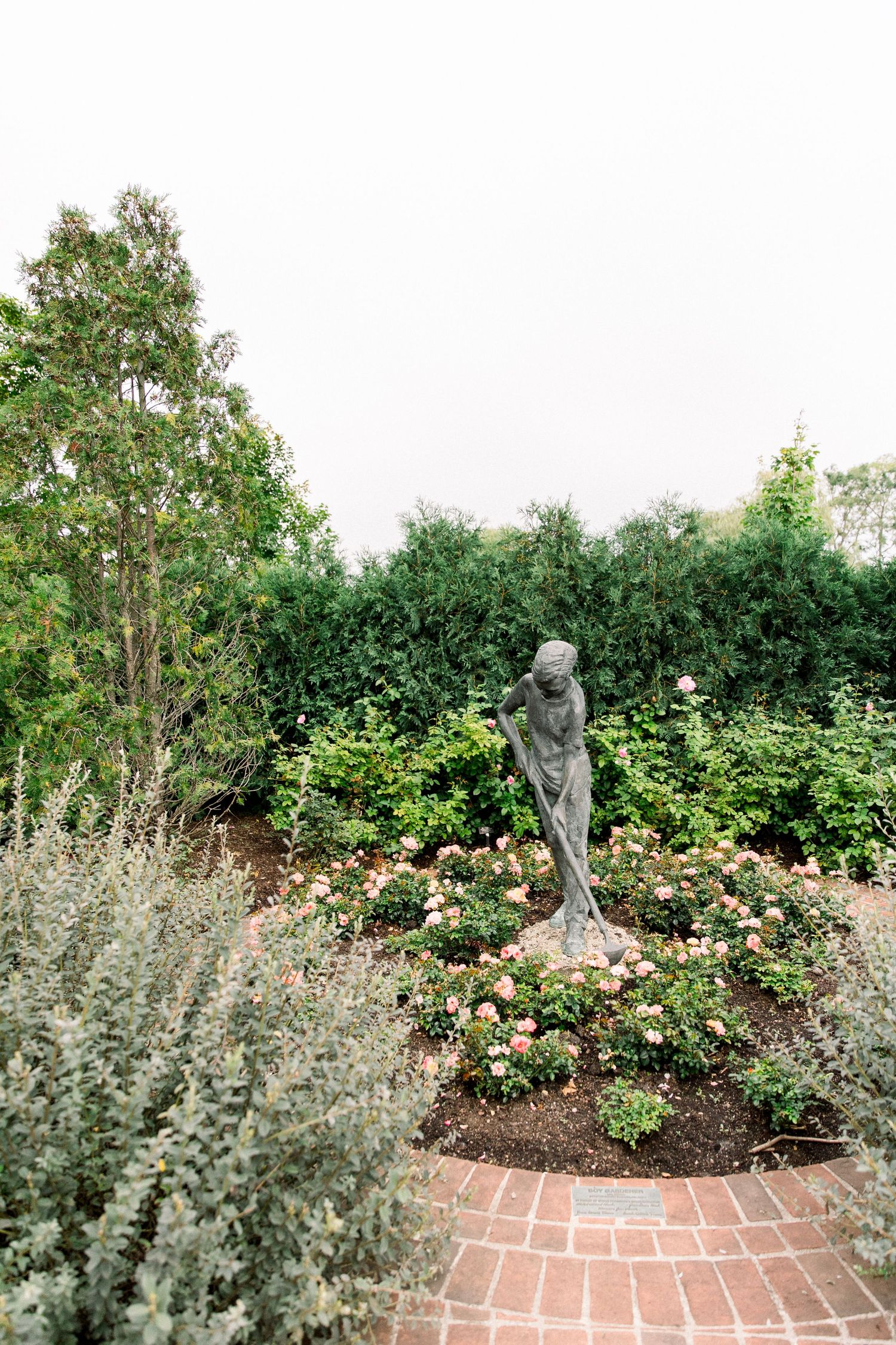 A bronze female statue surrounded by pink roses and lush green hedges in a formal garden with brick pathways.