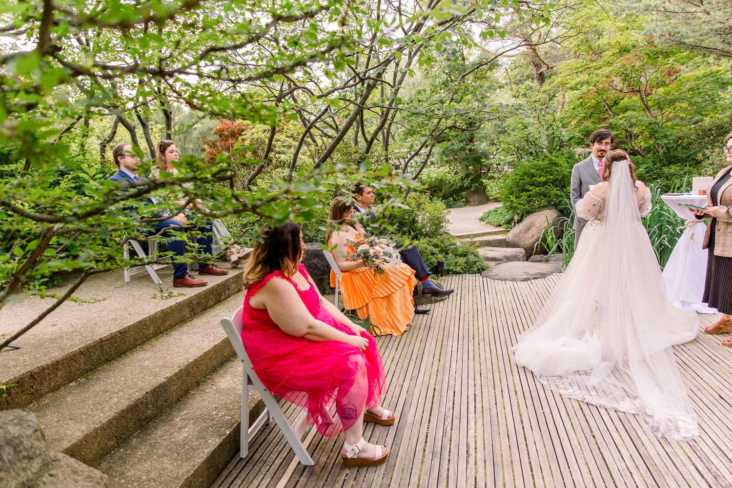 Bride and groom share a kiss on a wooden deck during an outdoor garden wedding ceremony with guests seated nearby.
