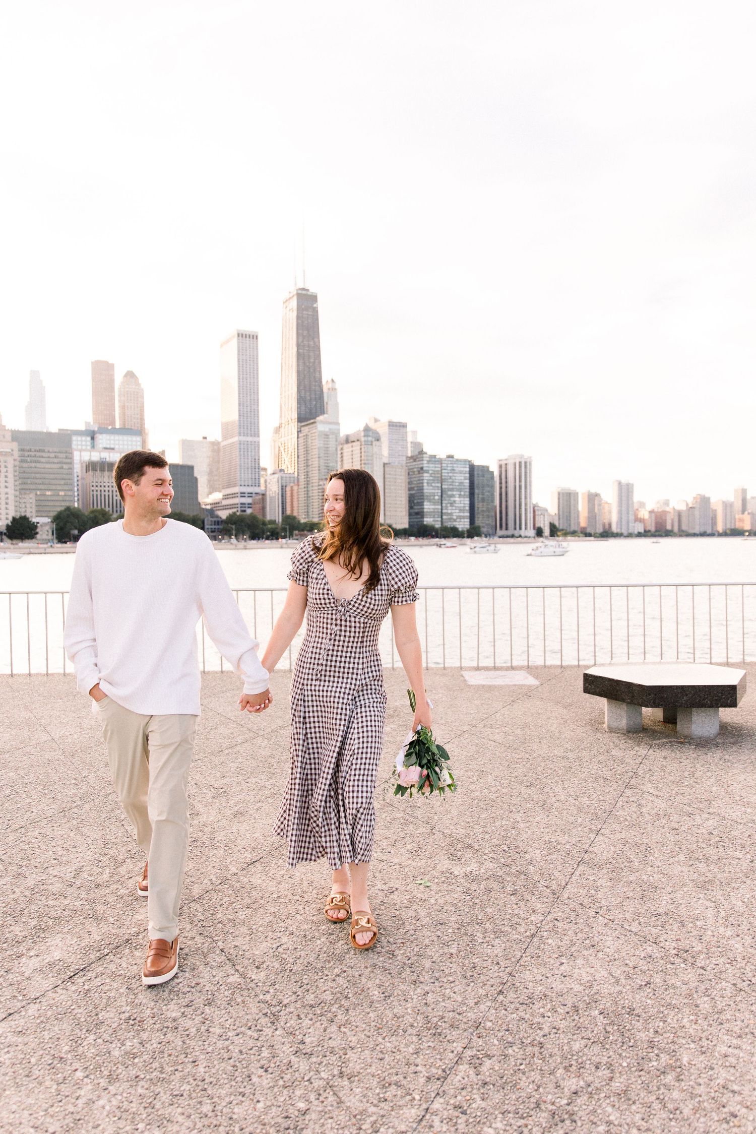Couple holding hands and walking along Chicago waterfront, woman in gingham dress holding flowers with city skyline behind them.