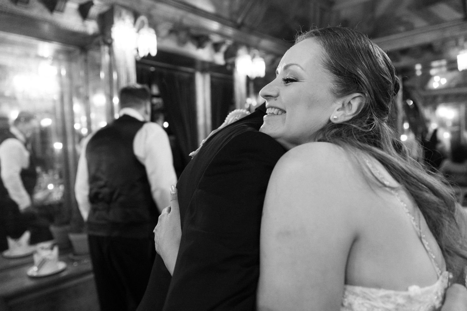 Black and white photo of a smiling bride embracing a guest in formal wear at an elegant wedding reception.