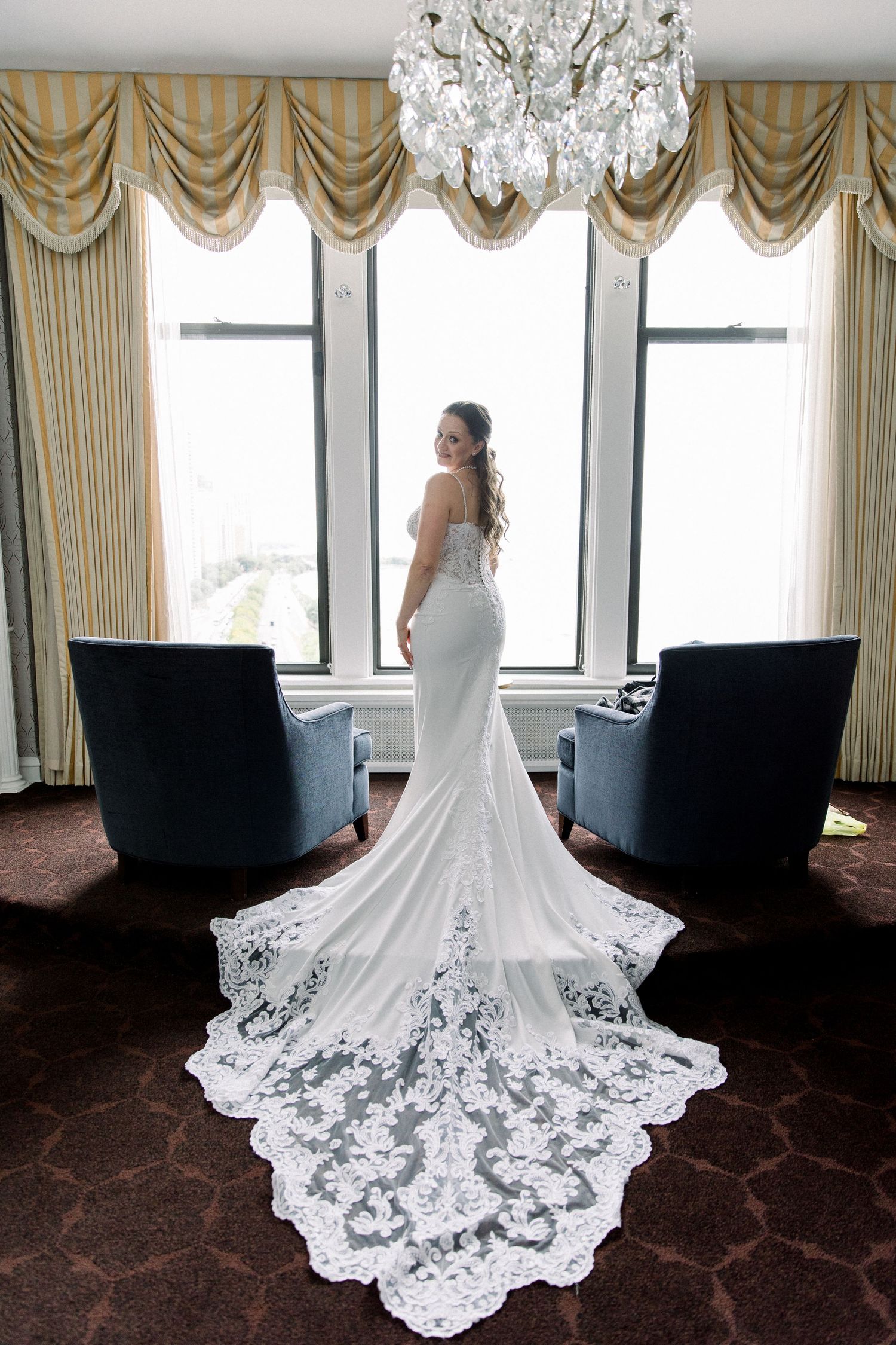 Bride in elegant lace train wedding gown standing by large window with chandelier above.