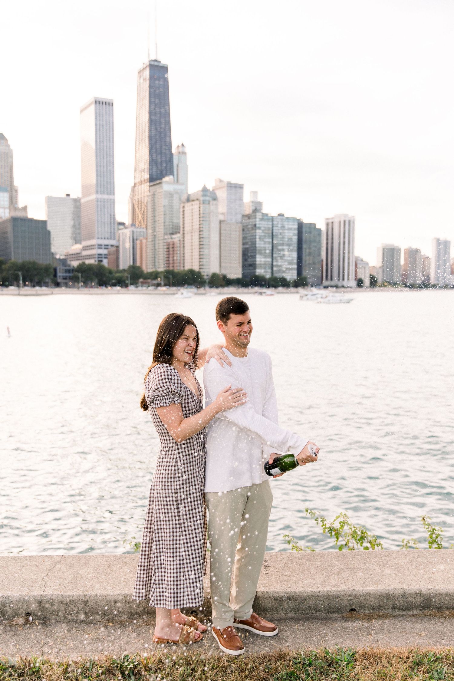 Couple celebrating with champagne by Chicago lakefront skyline, woman in gingham dress, man in white shirt, sunny day.