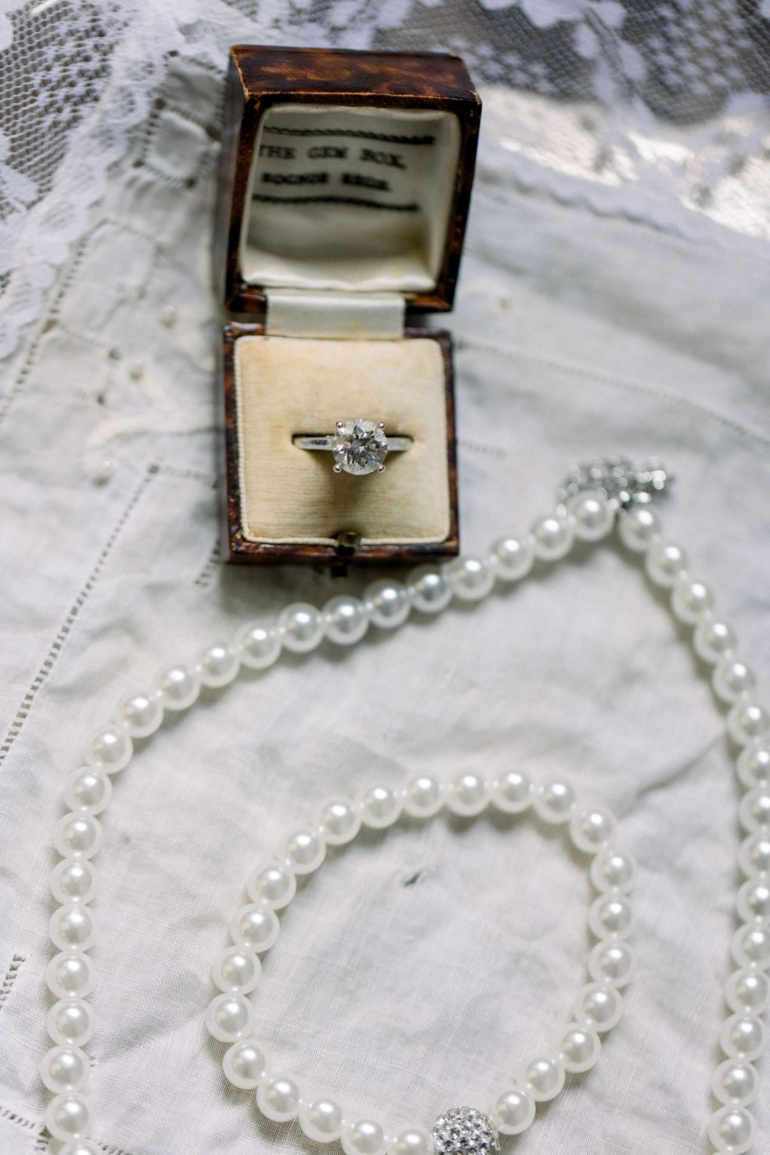Close-up of diamond solitaire ring in antique jewelry box beside a pearl necklace on white lace.