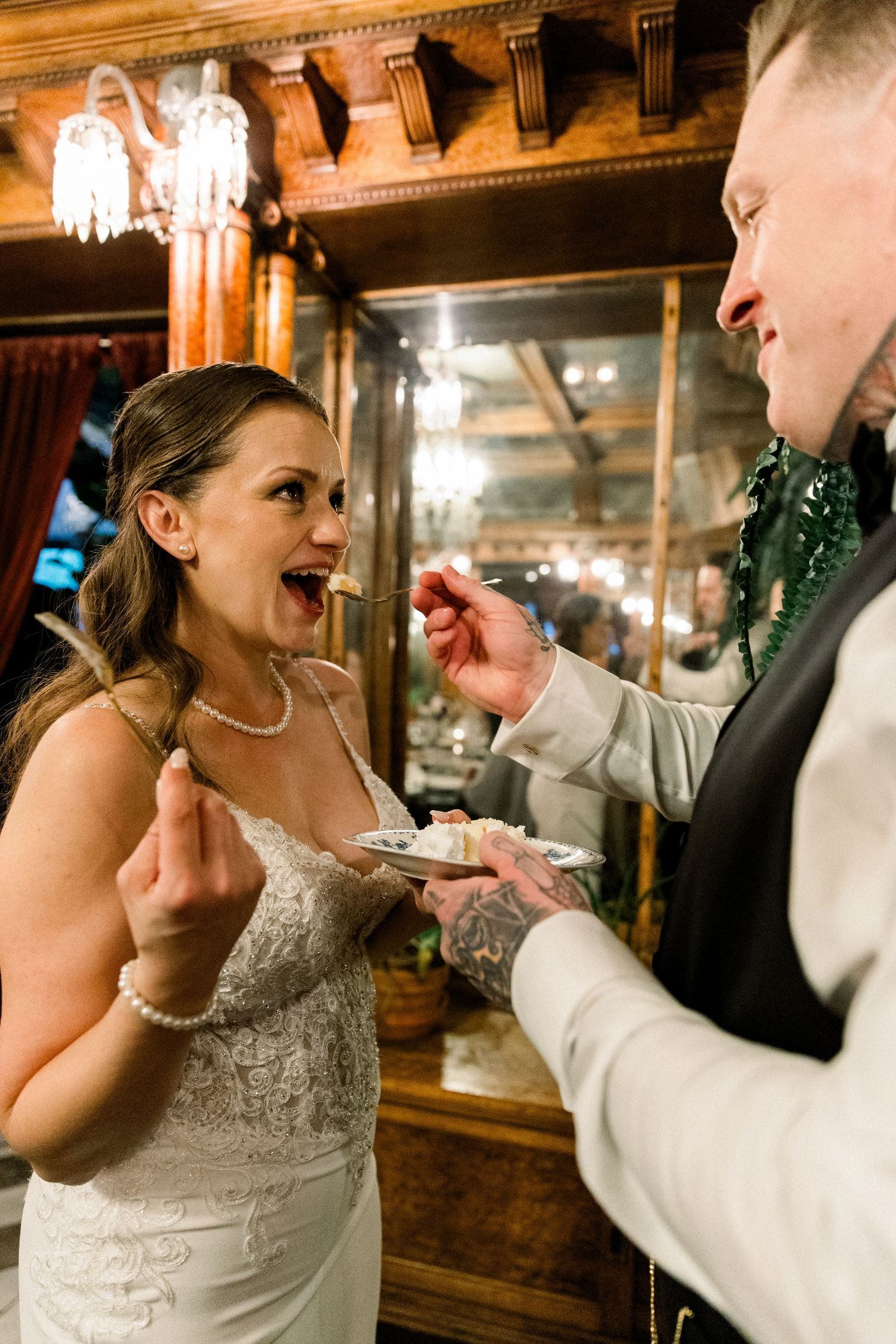 Groom feeds bride a bite of wedding cake in an elegant warmly lit reception venue.