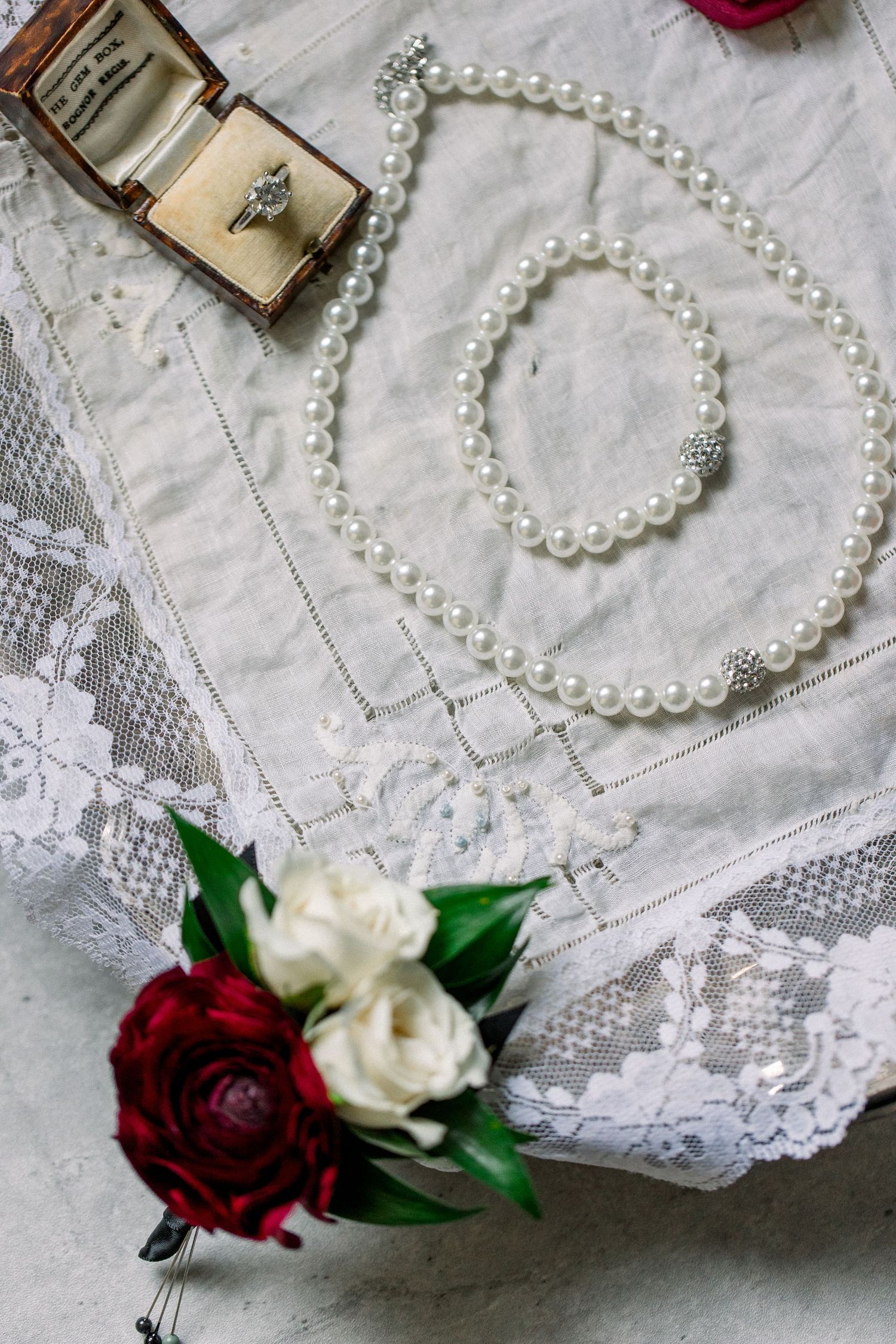 Pearl necklace, diamond engagement ring in vintage box, and red and white floral bouquet on lace fabric.