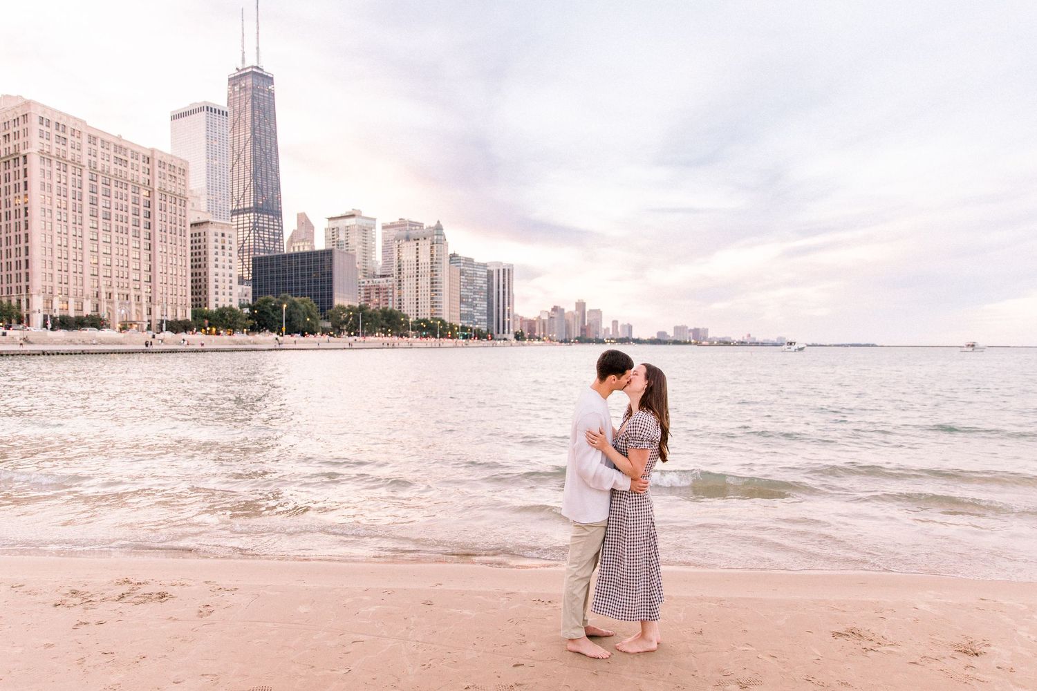 Couple kissing on Chicago beach with city skyline and John Hancock Center in background at sunset.