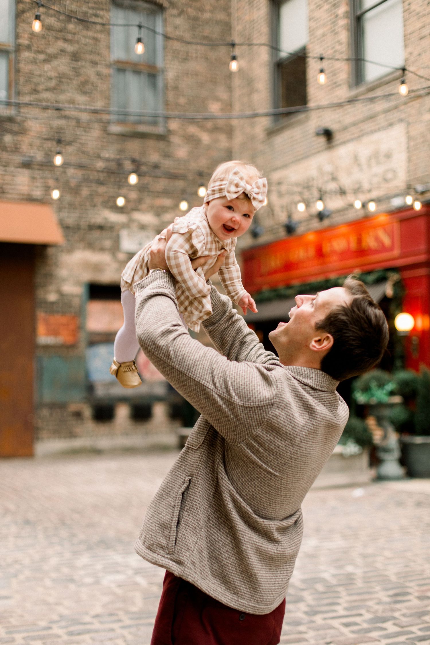 Father joyfully lifting smiling baby in checkered outfit outdoors near brick building with string lights.