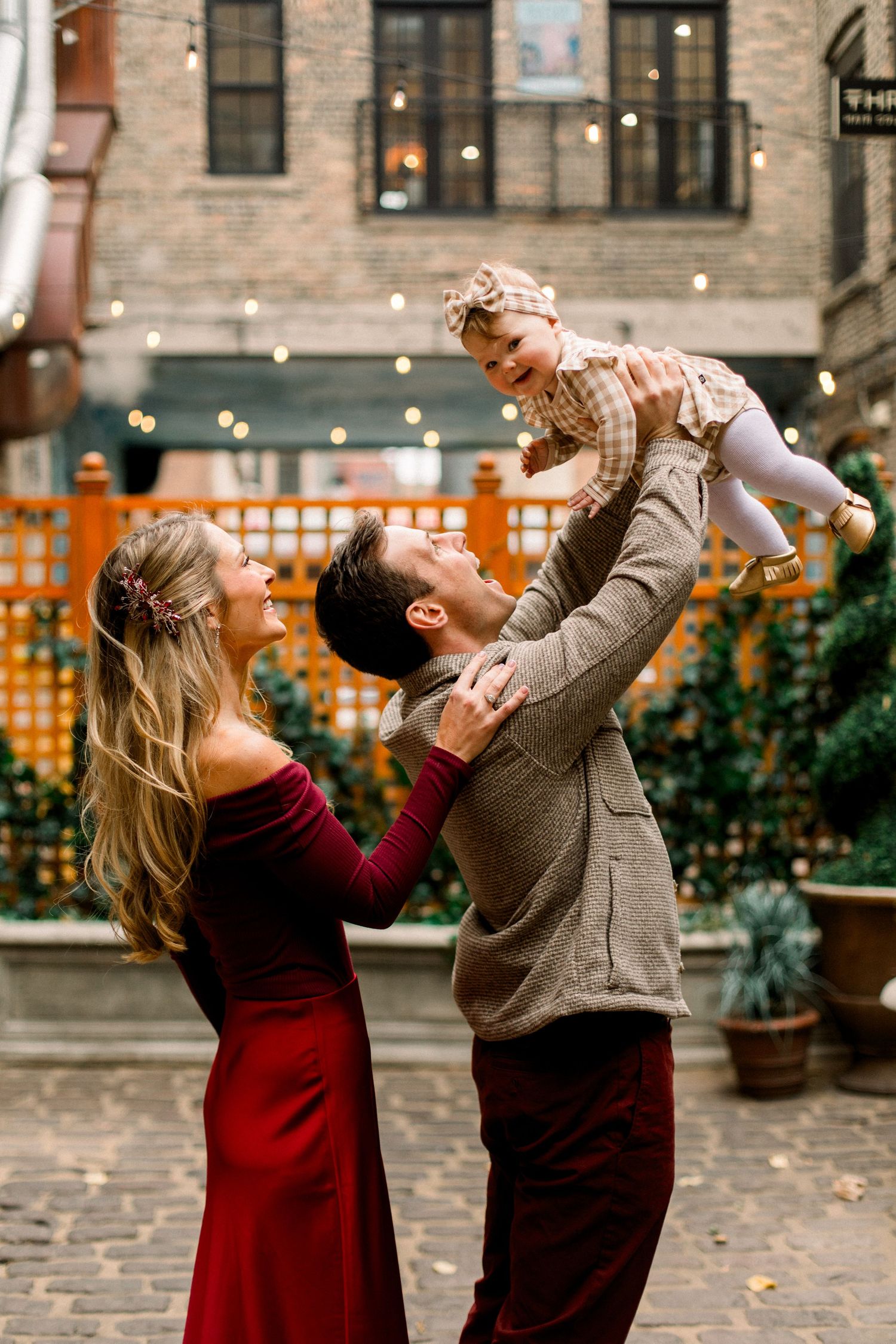 A joyful family poses outdoors, with a man lifting a baby girl while a woman in a red dress smiles beside them.
